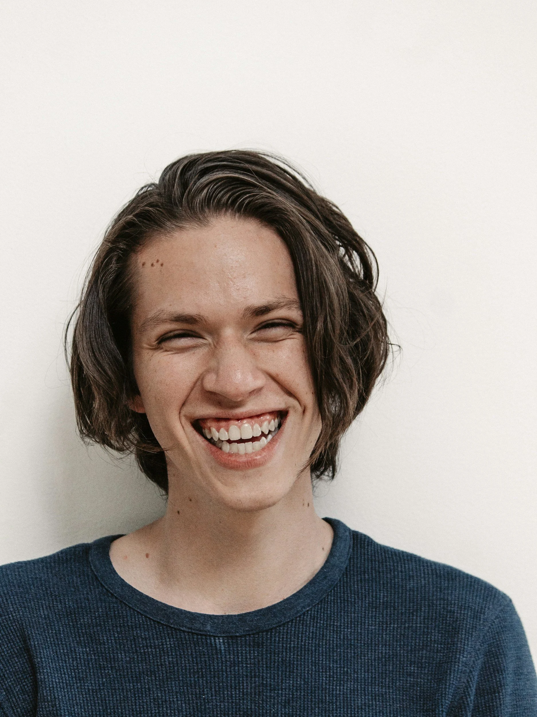 A young woman with short, dark brown, slightly wavy hair, smiling with eyes nearly closed, wearing a dark blue textured shirt, against a plain white background.