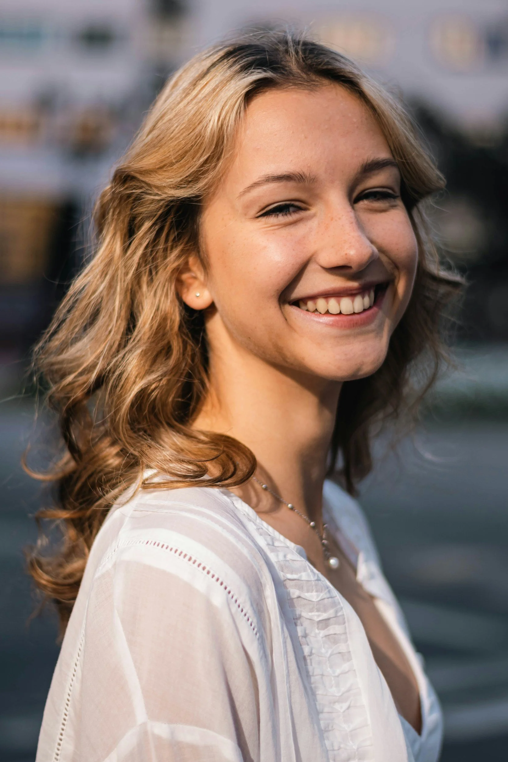Close-up portrait of a young woman with wavy, light brown hair smiling outdoors near water, wearing a white top with lace details and a pearl necklace.