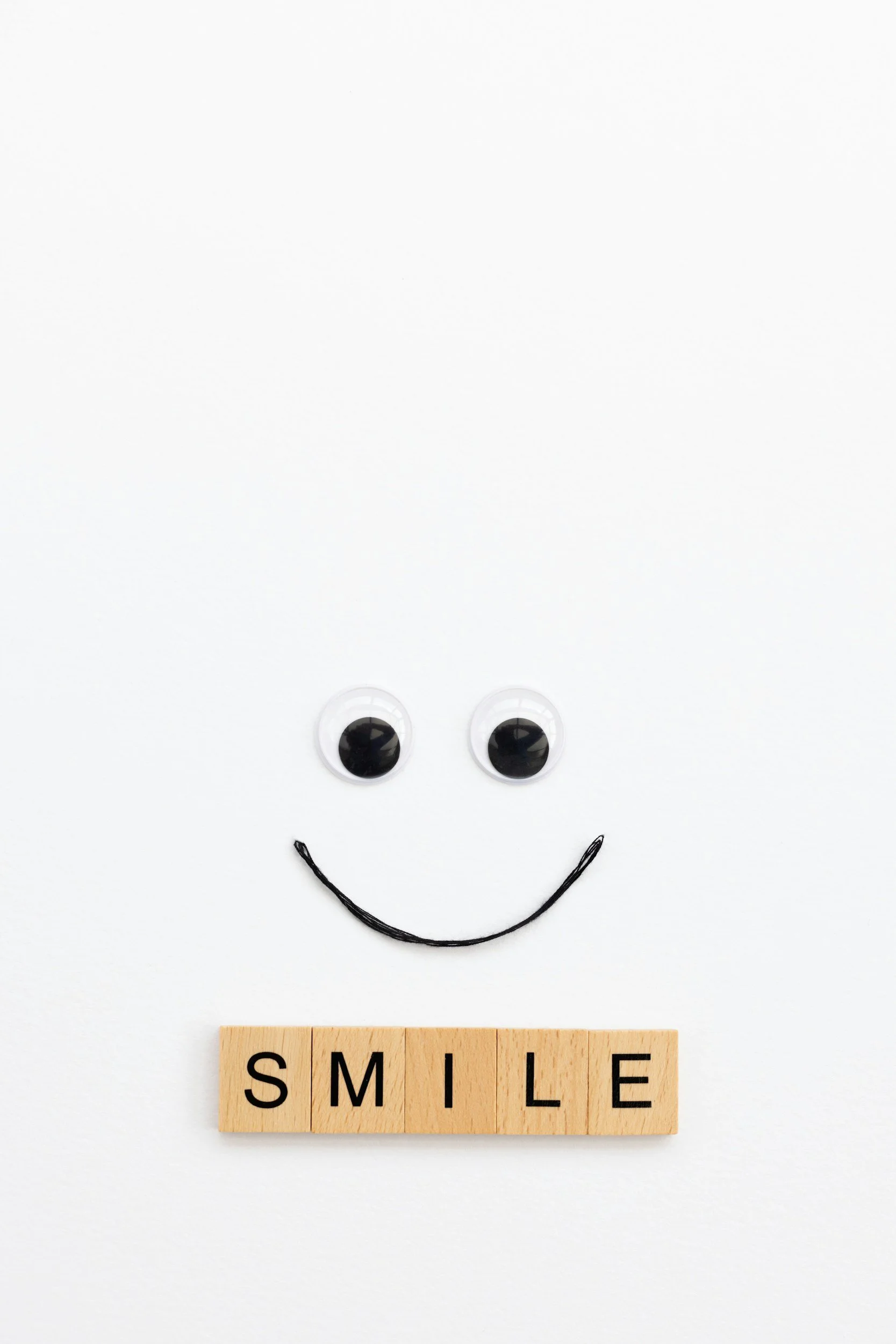 A white background with a smiley face made from googly eyes for eyes, a small black yarn for a curved smile, and a light wood block with the word 'SMILE' spelled out in black letters at the bottom.