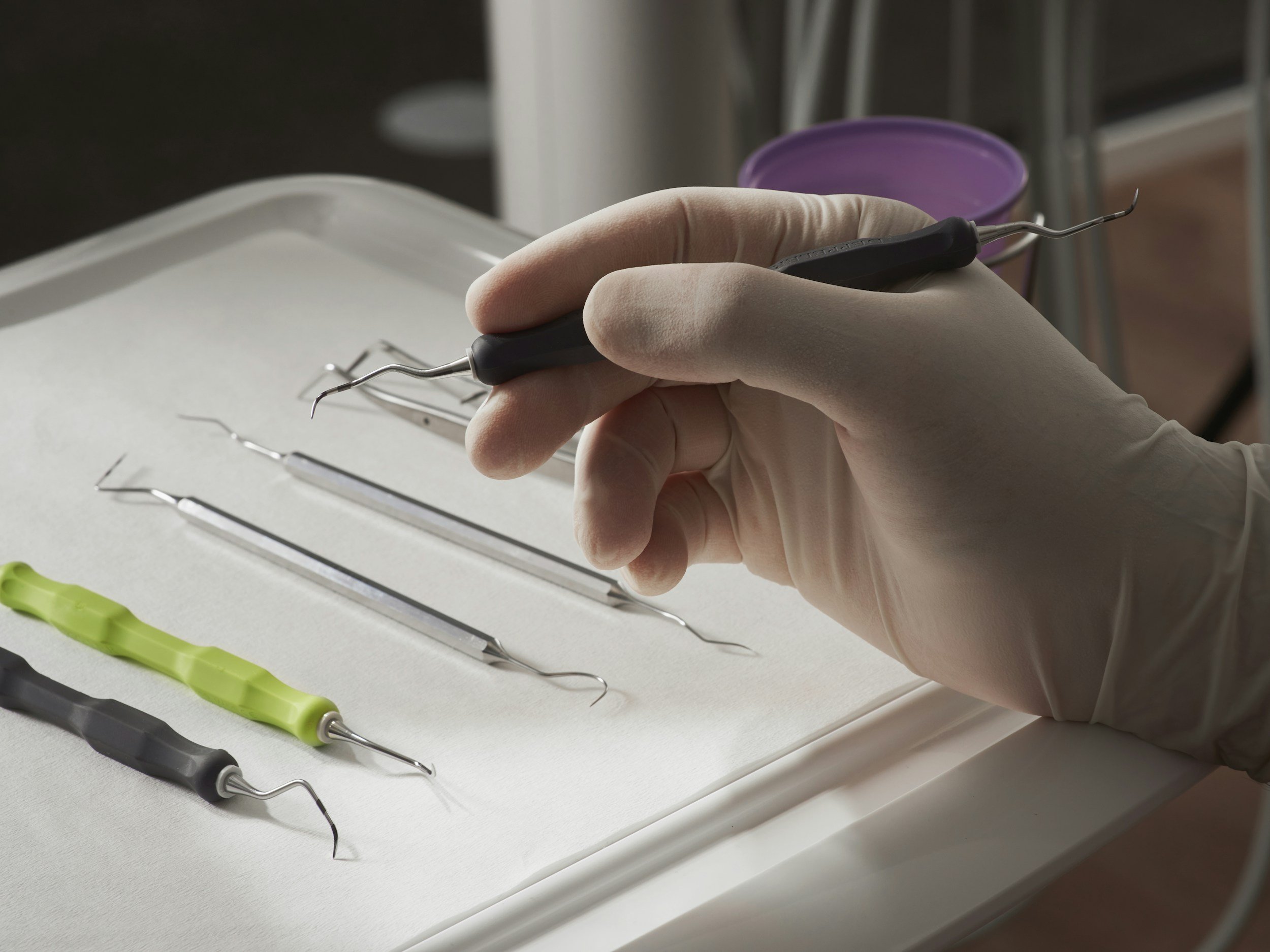 A gloved hand holding a dental or surgical tool with a curved metal tip, placed on a white surface with other similar tools nearby.