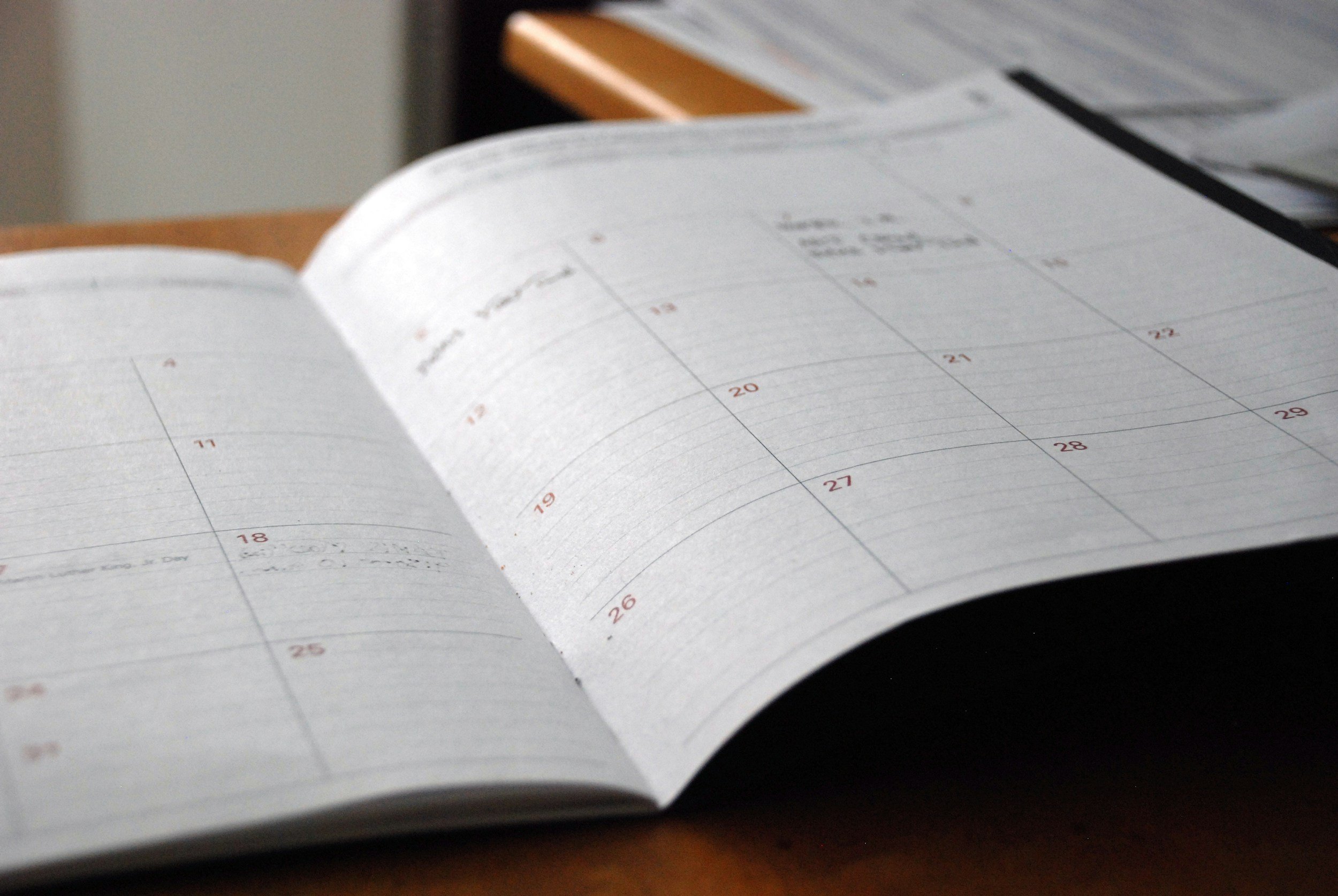An open planner or calendar laid out on a wooden table, displaying dates and days of the week, with a black pen and some documents in the background.