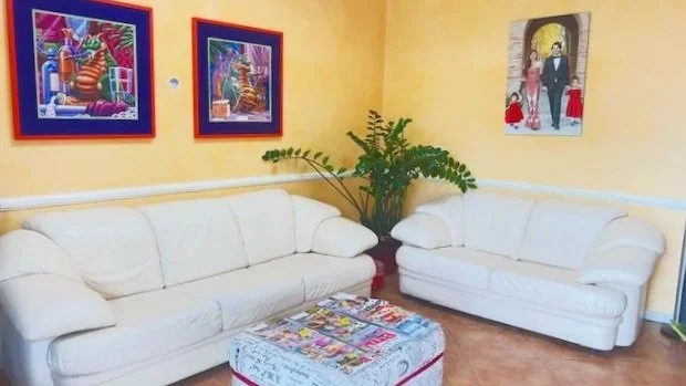 Living room with two white sofas, colorful artwork on yellow walls, a large green potted plant, and a patterned ottoman in the center.