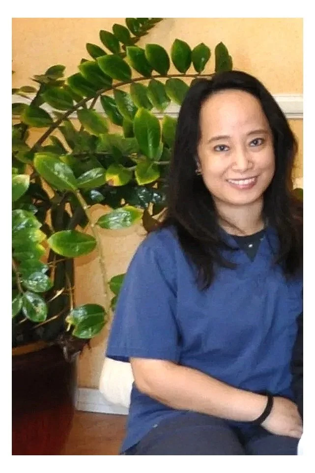 A woman with dark hair smiling, wearing a blue shirt, sitting indoors next to a large green leafy plant.