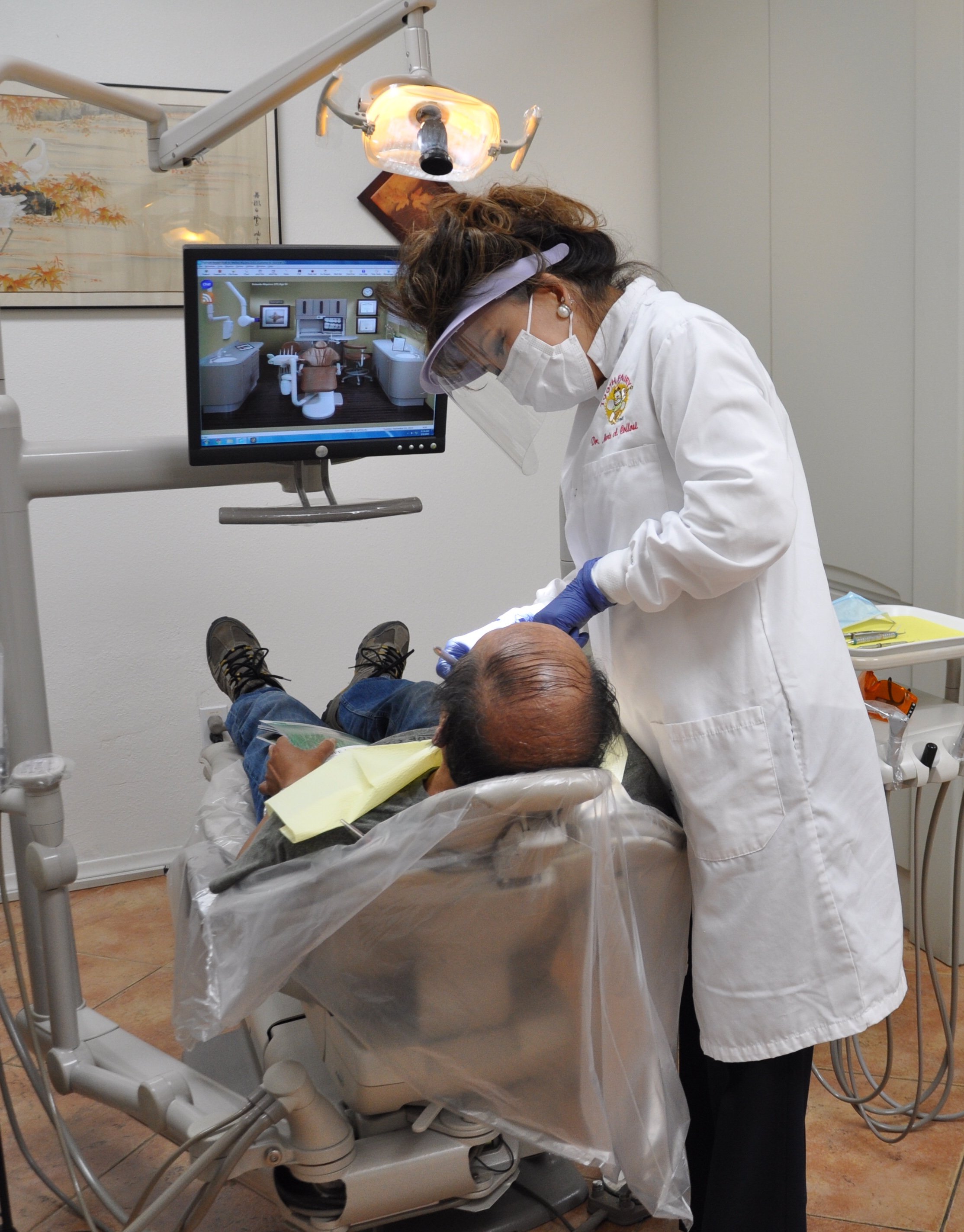 A dentist in a white coat, gloves, and mask working on a patient in a dental chair. The dentist wears a face shield and is looking down at the patient, who is lying back with a yellow dental bib. A computer monitor displays a virtual dental office in the background.