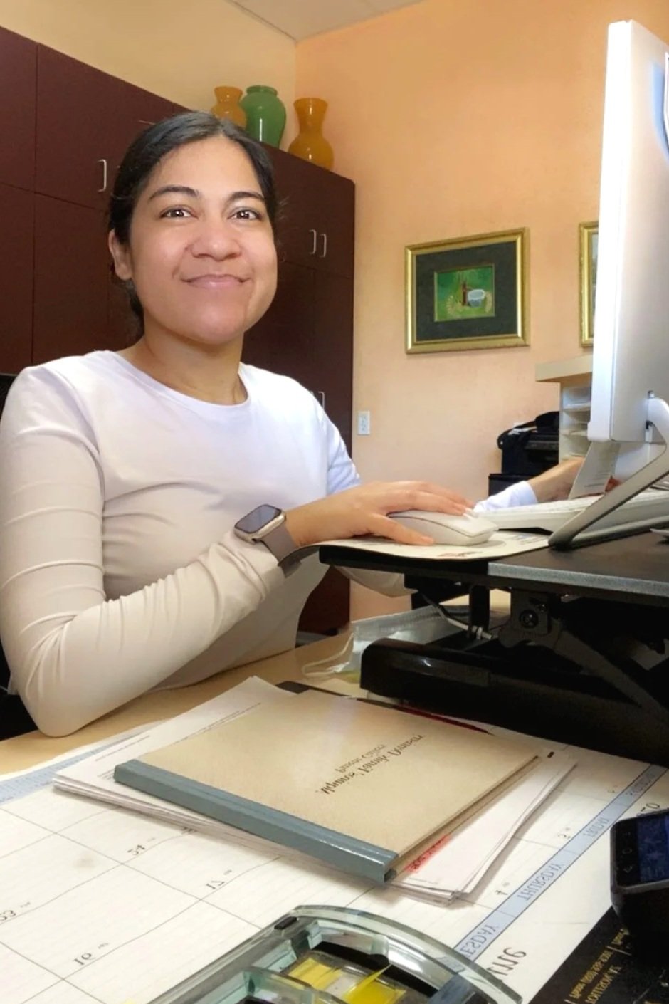 A woman sitting at a desk with a computer, smiling at the camera, in an office setting with framed pictures and decorative vases in the background.