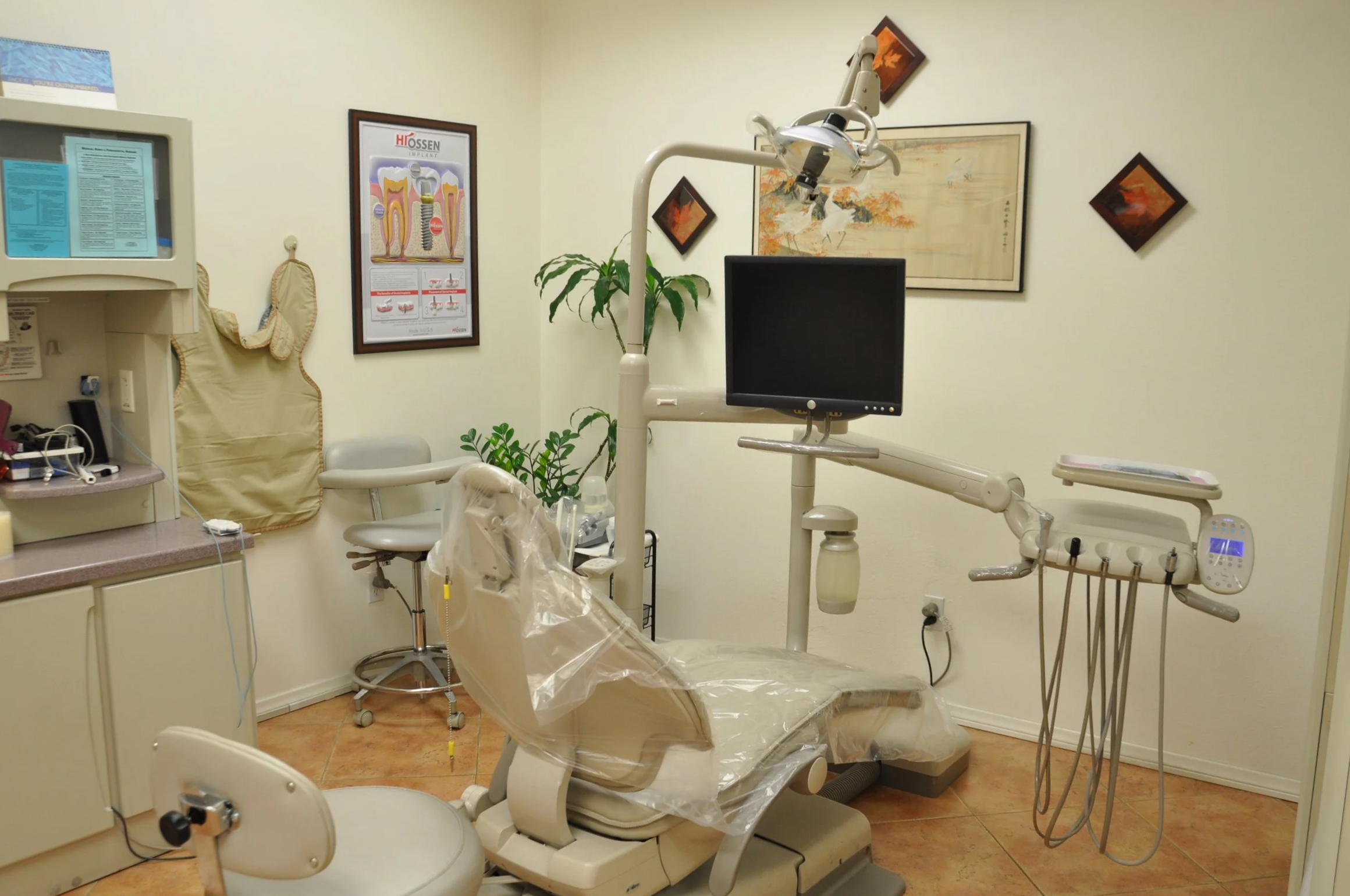 An empty dental examination room with a dental chair covered in a clear plastic protective sheet, surrounded by various dental equipment including a monitor, dental tools, and an overhead light, with framed artwork and posters on the walls.