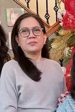 A woman with long dark hair, glasses, and a gray top standing indoors near festive decorations.