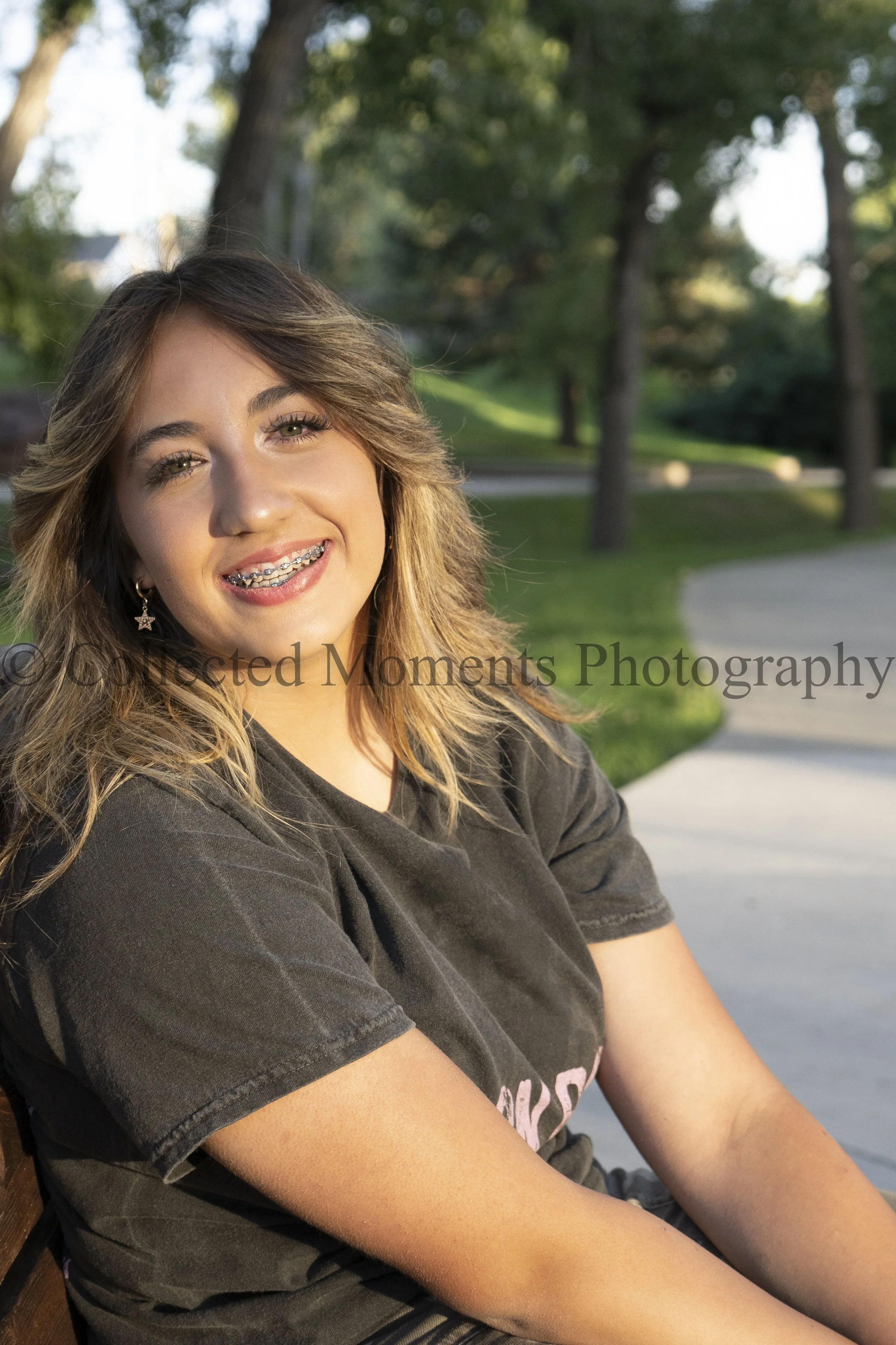 A young woman with light skin, long wavy brown hair with blonde highlights, and braces smiling outdoors in a park during daytime. She is wearing star-shaped earrings and a dark T-shirt. The background features green trees, a grassy area, and a sidewalk.