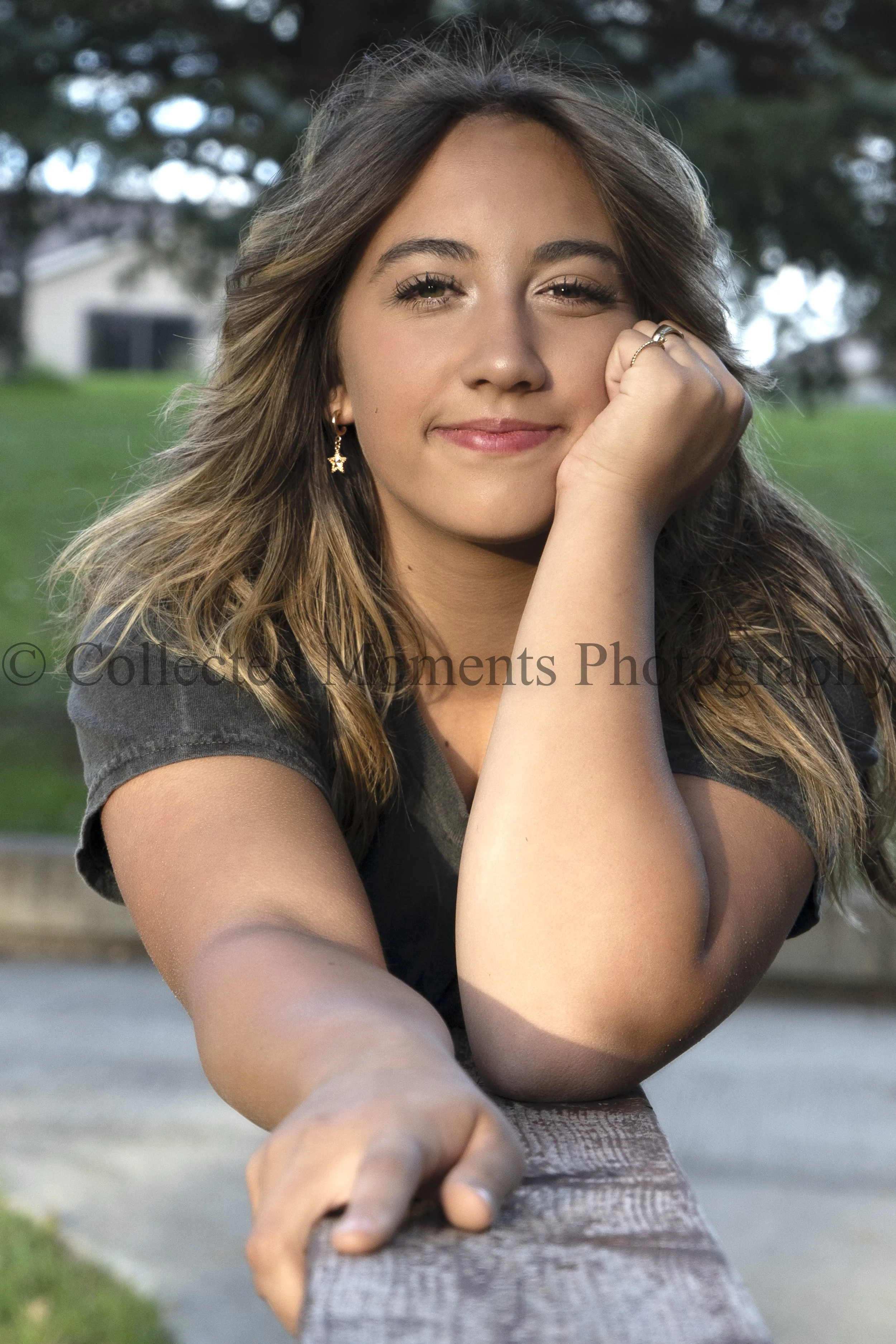 Close-up of a young woman smiling, with her chin resting on her hand, outdoors in a park-like setting with trees and a building in the background.