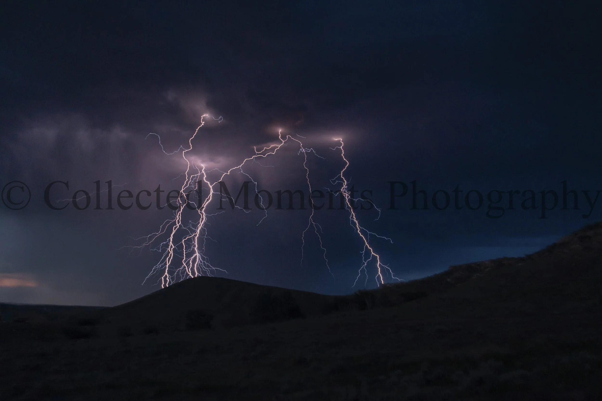 Lightning strikes illuminating a dark stormy sky over a mountainous landscape at night.
