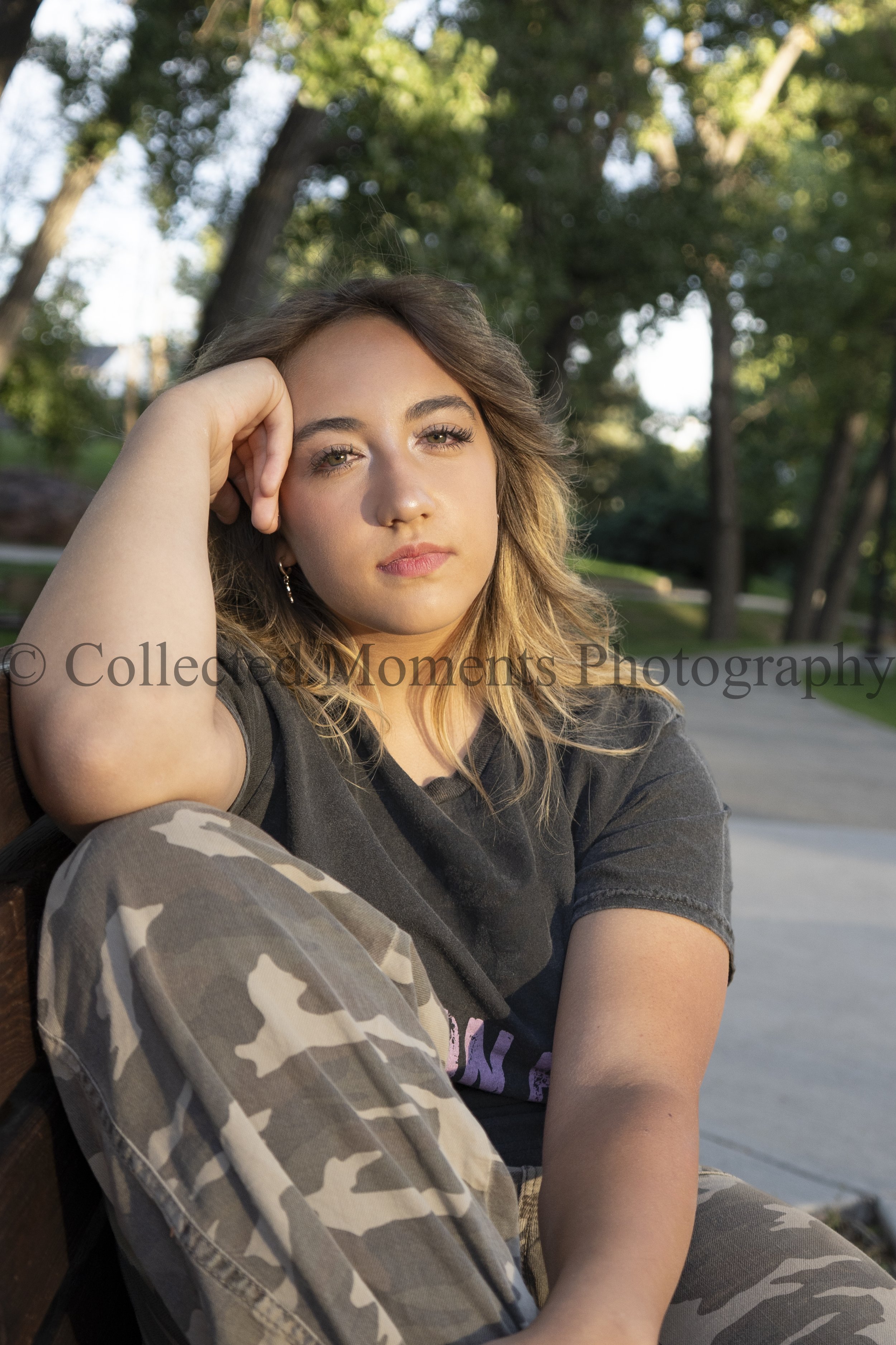 A young woman with long wavy hair, wearing a black t-shirt and camo pants, sitting outdoors near a park bench with trees in the background, resting her head on her hand.