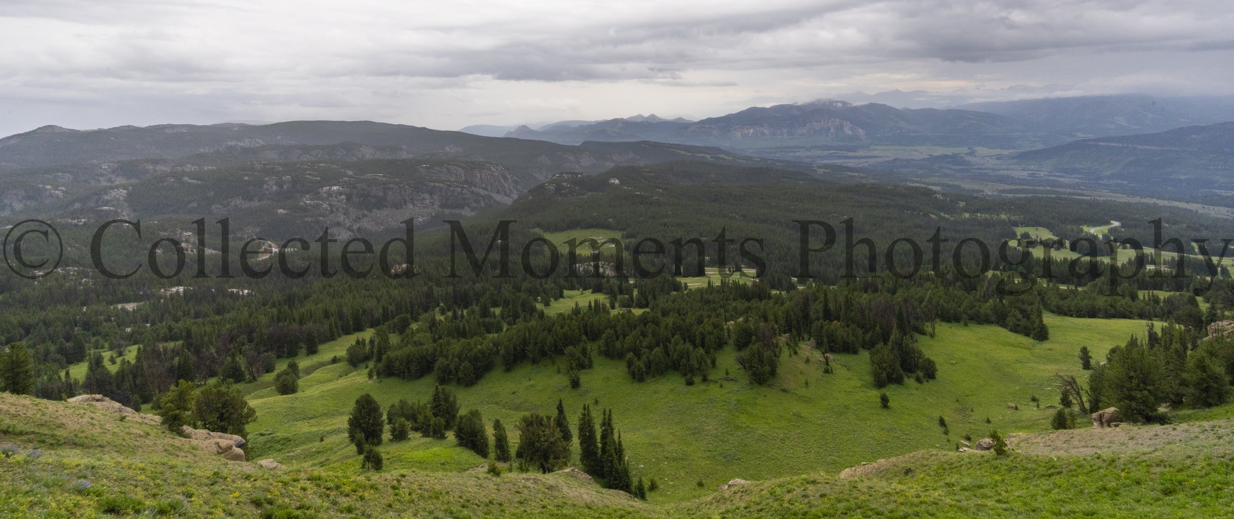 Scenic view of green hills, forest, and distant mountains under cloudy sky in a rural landscape.
