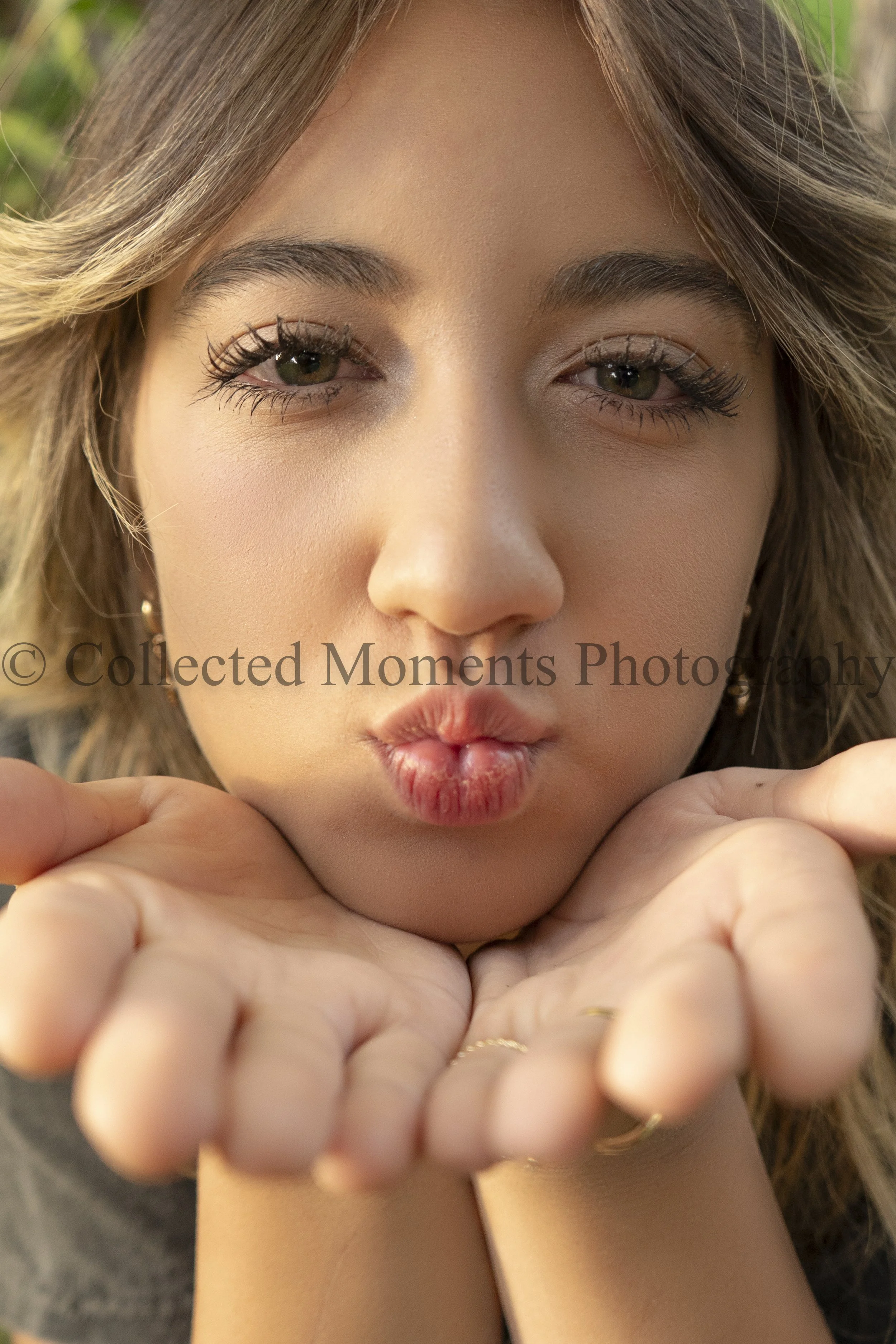 Close-up of a woman's face with her hands under her chin, making a kissing face. She has light makeup, brown eyes, long eyelashes, and wavy light brown hair. There is sunlight highlighting her features.