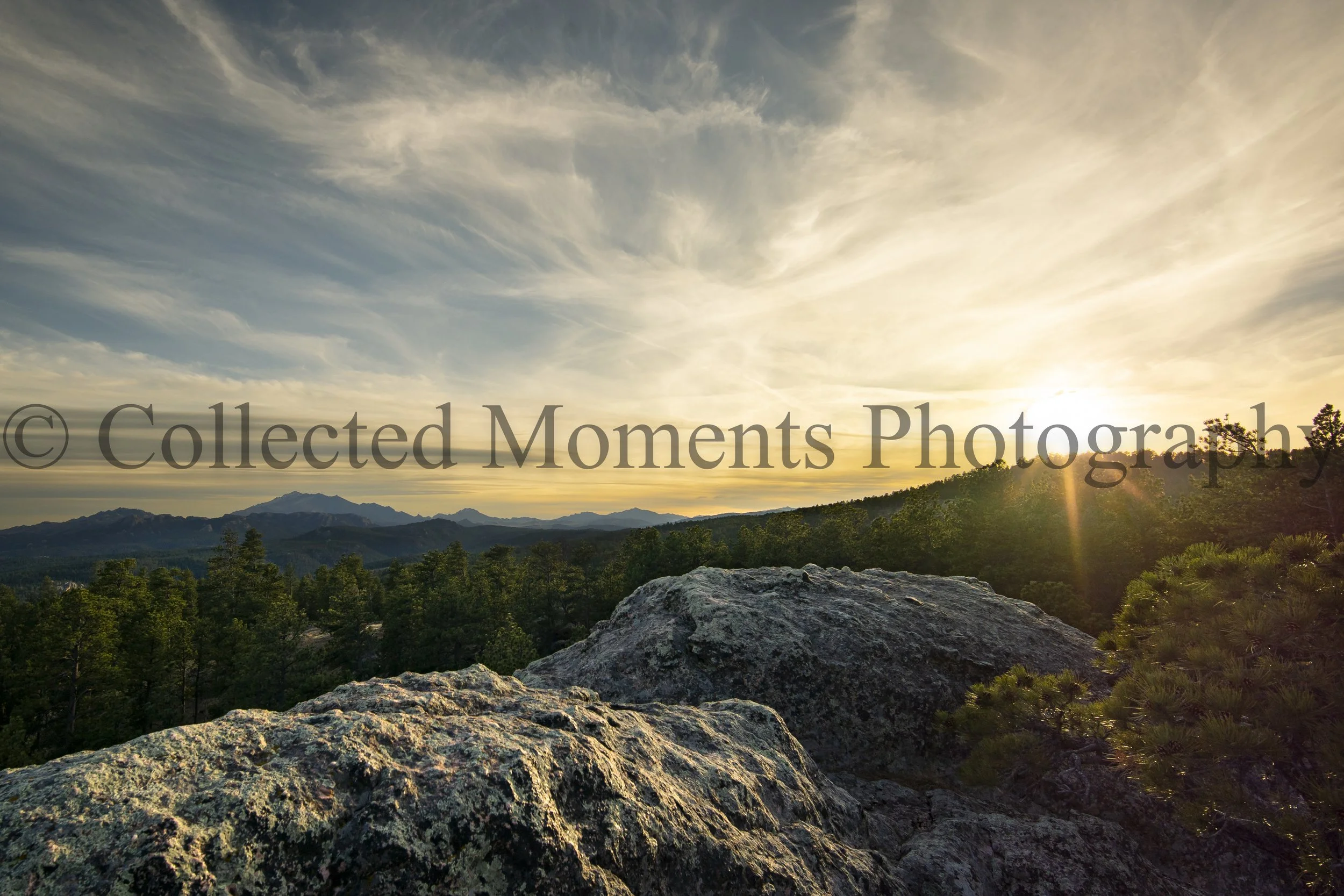 Sunset over a mountainous landscape with trees in the foreground and a large rock in the lower center.