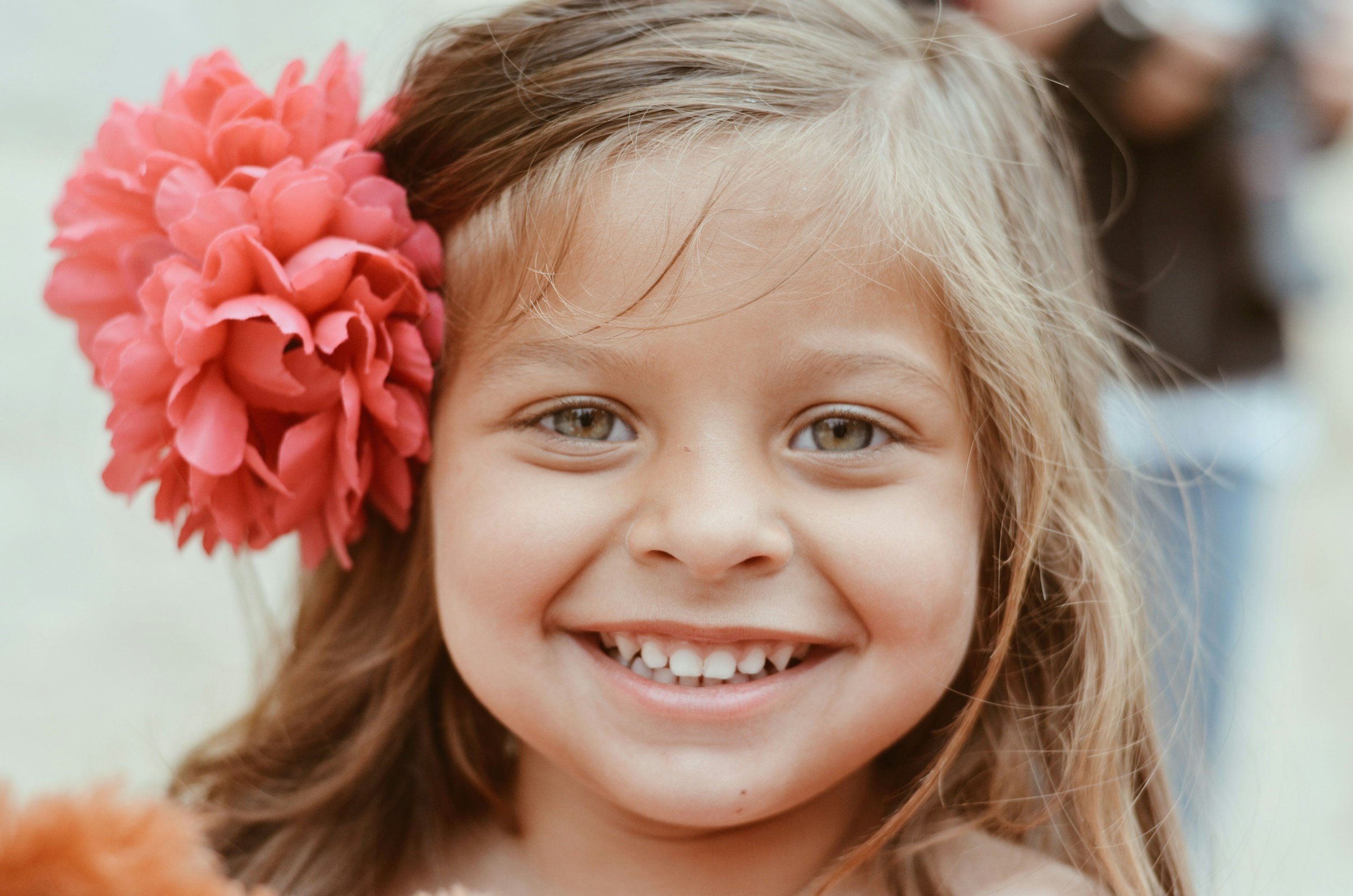 Close-up of a young girl smiling, with light brown hair, hazel eyes, and a large pink flower in her hair.