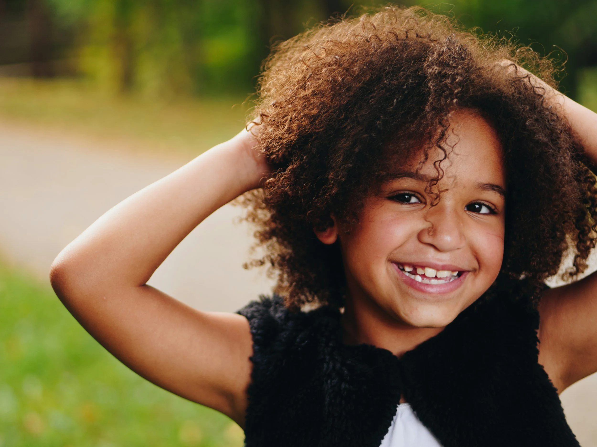 Close-up of a young girl with curly brown hair smiling outdoors, with blurred green and yellow background.