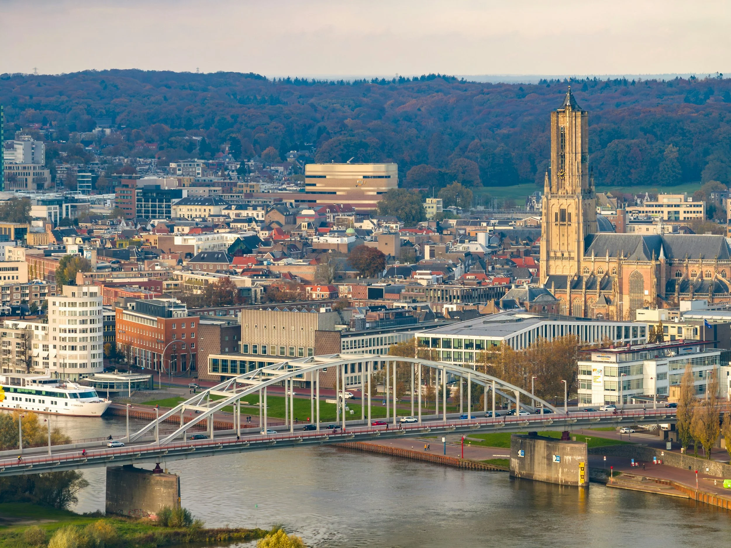 A cityscape of Arnhem featuring a river in the foreground, a bridge crossing over the river, and various modern and historic buildings in the background, with a hill covered in trees in the distance.