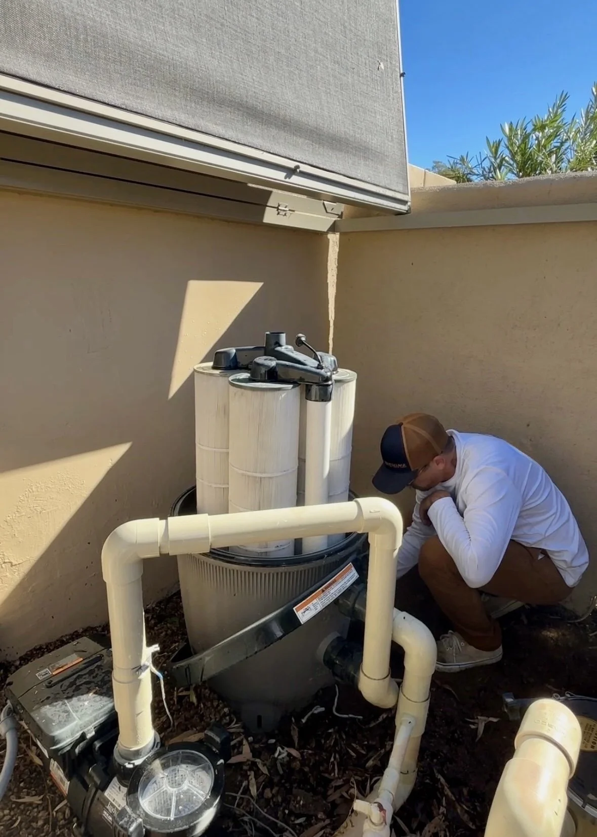 A man wearing a white shirt, brown shorts, and a brown cap crouches beside a pool filtration system with white pipes and a large filter tank. He appears to be working on or inspecting the equipment outdoors, with a beige stucco wall and a blue sky in