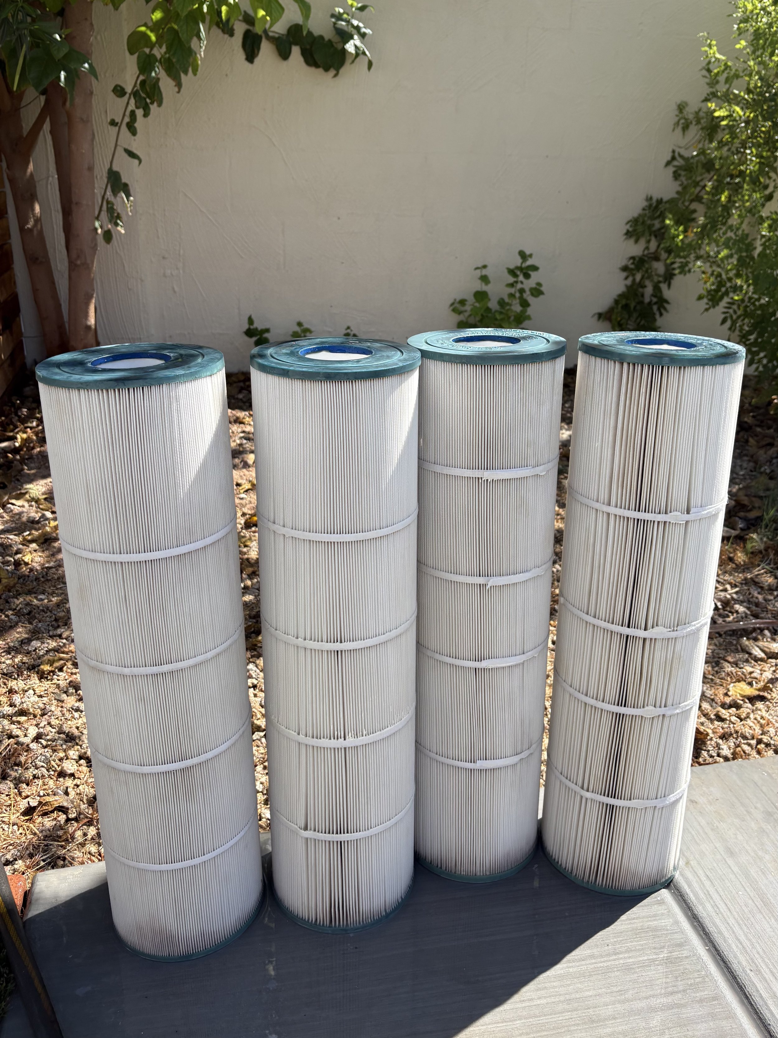 Four white pool filters with blue tops placed outdoors on a gray surface, with a white wall, green leafy plants, and trees in the background.