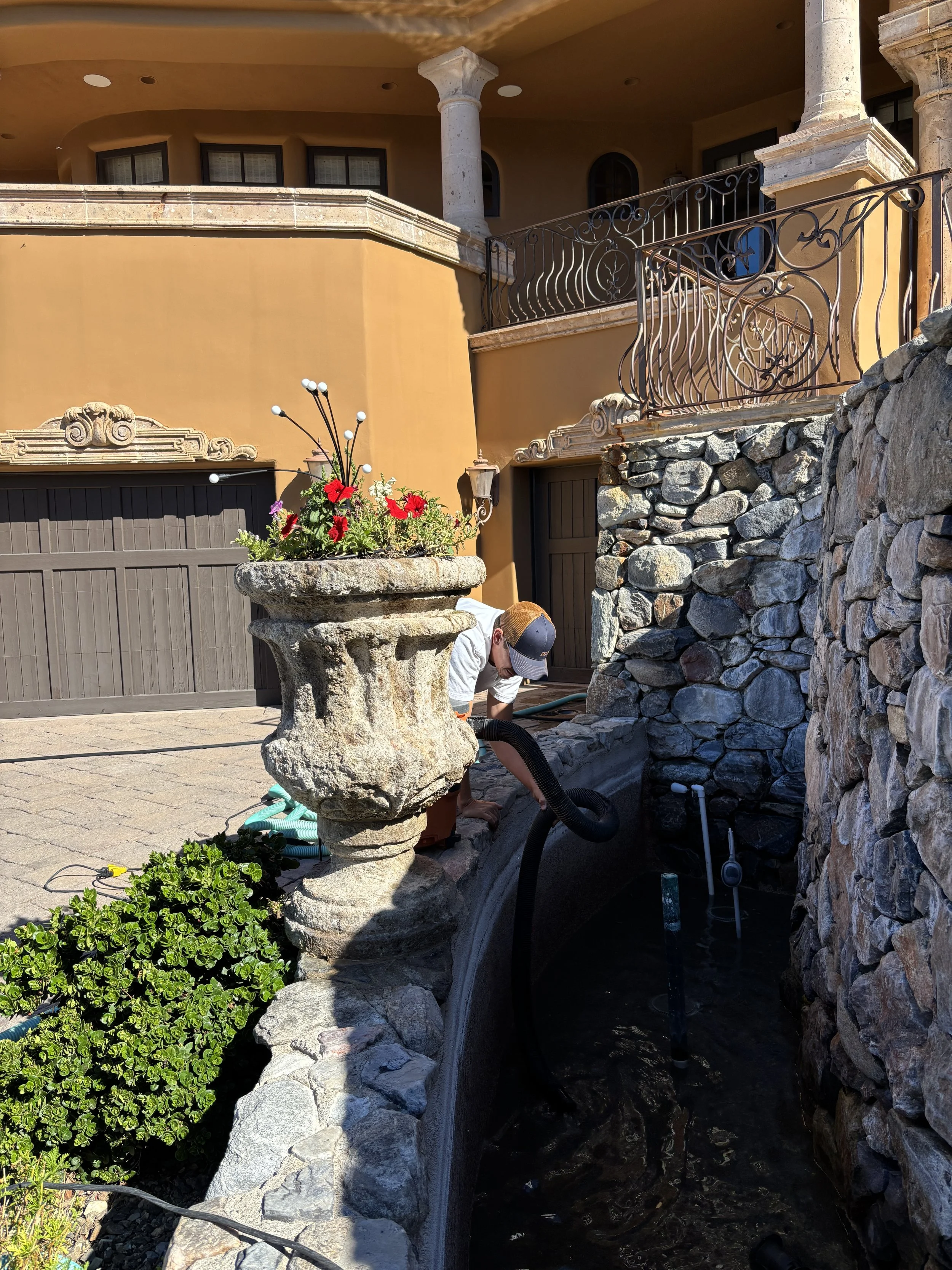 A man wearing a cap and white T-shirt working on a stone and brick fountain in a courtyard with a yellow building, stone walls, a flower planter, and decorative metal railings.