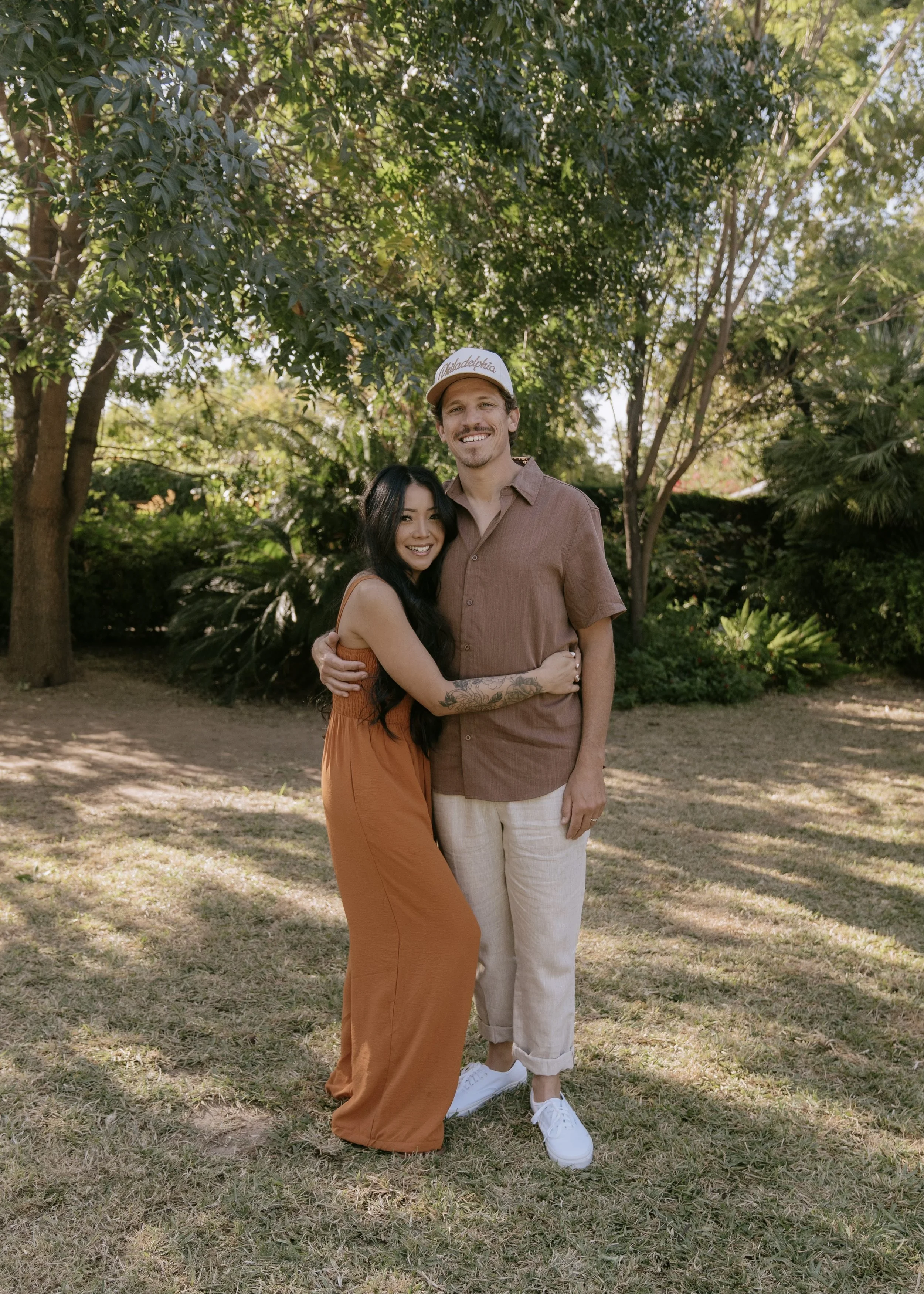 Two people, a woman with long dark hair and a man with light hair and a beard, are standing outdoors under trees, hugging and smiling at the camera. The woman is wearing a burnt orange dress, and the man is wearing a brown shirt, beige pants, white sneakers, and a white cap that says "Philadelphia."