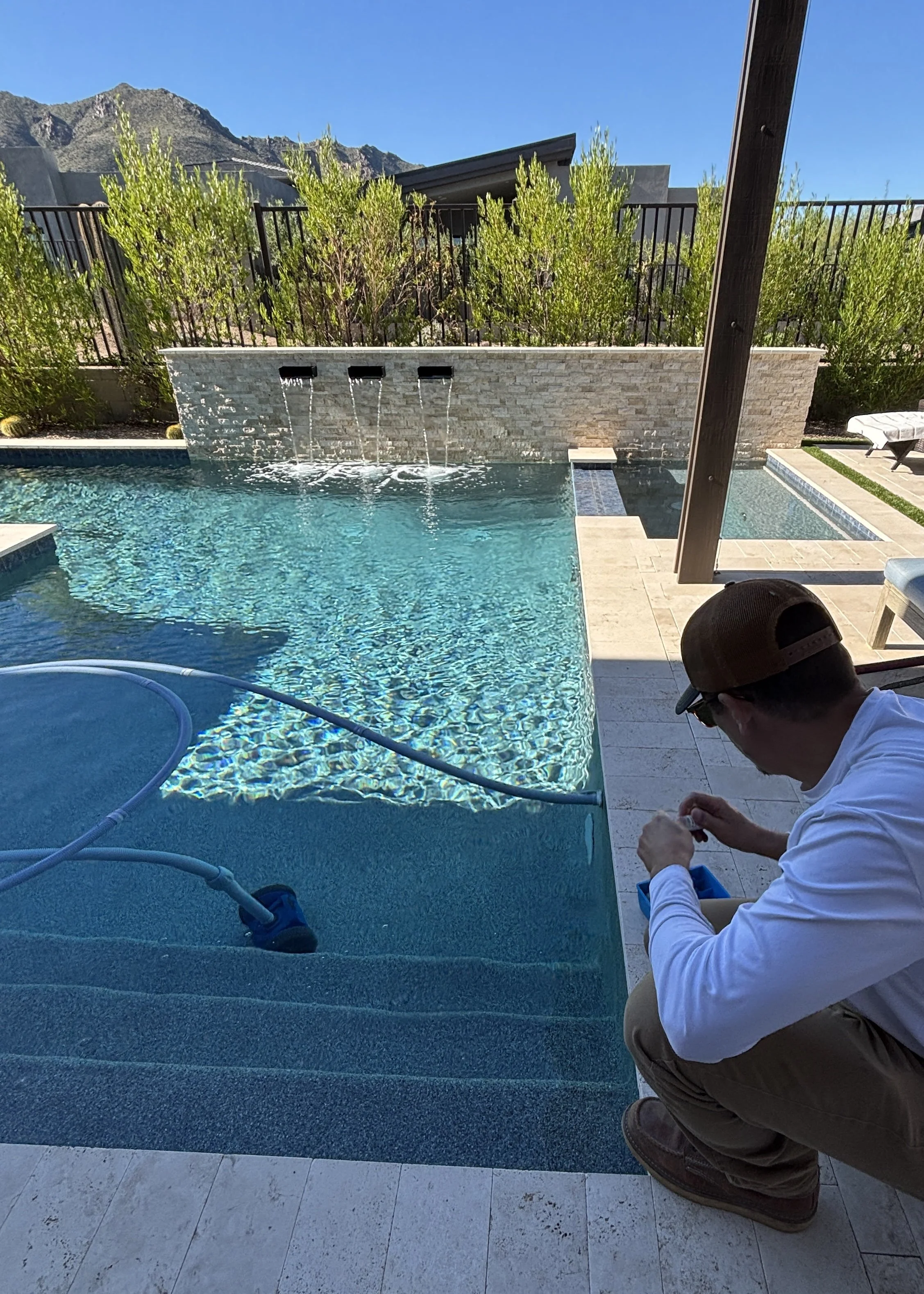 Man kneeling next to a swimming pool, working on cleaning or maintenance equipment, with a mountain and blue sky in the background.