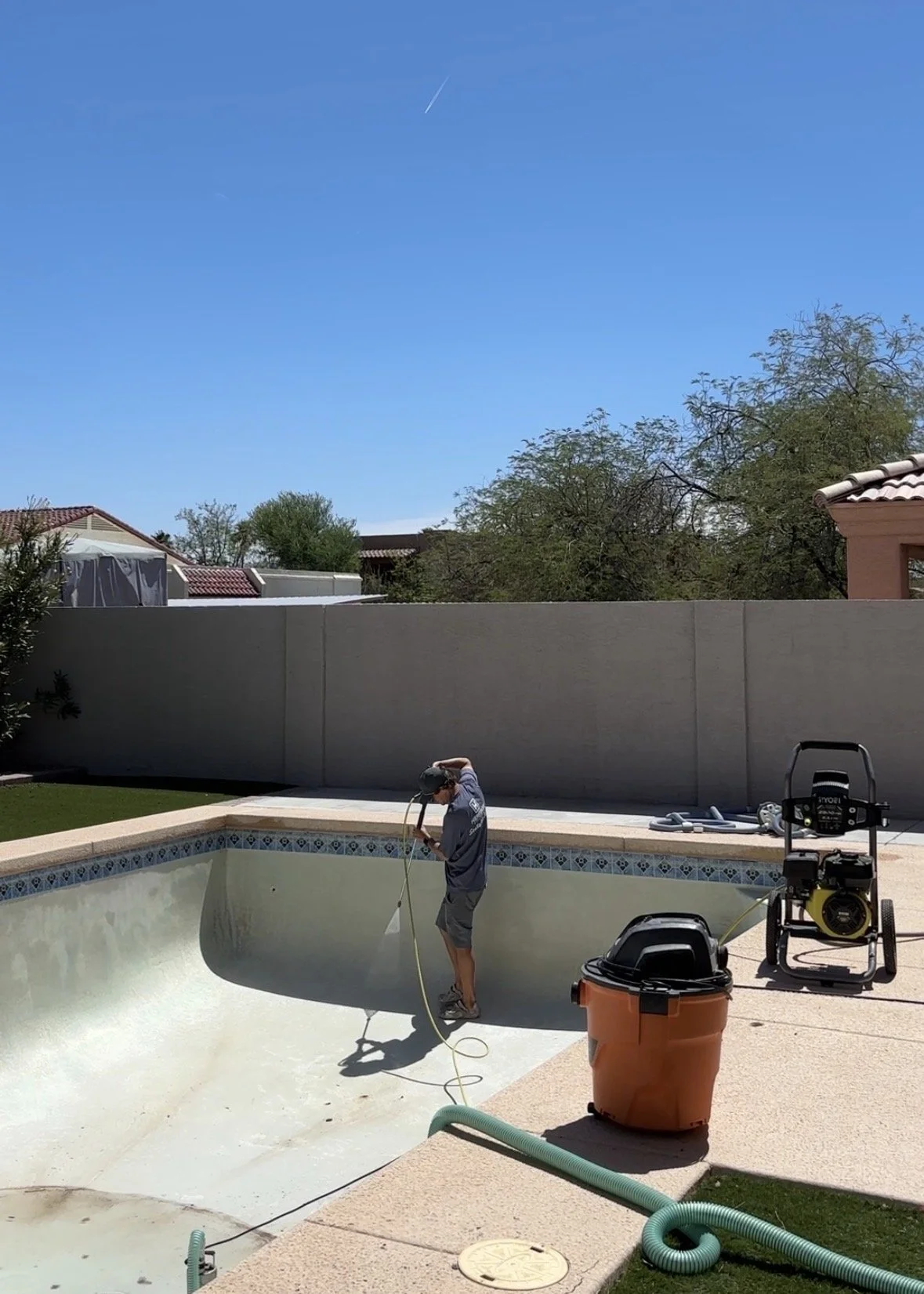 A person wearing a hat and gray shirt cleaning a backyard swimming pool using a hose, with pool cleaning equipment and a trash bin nearby.