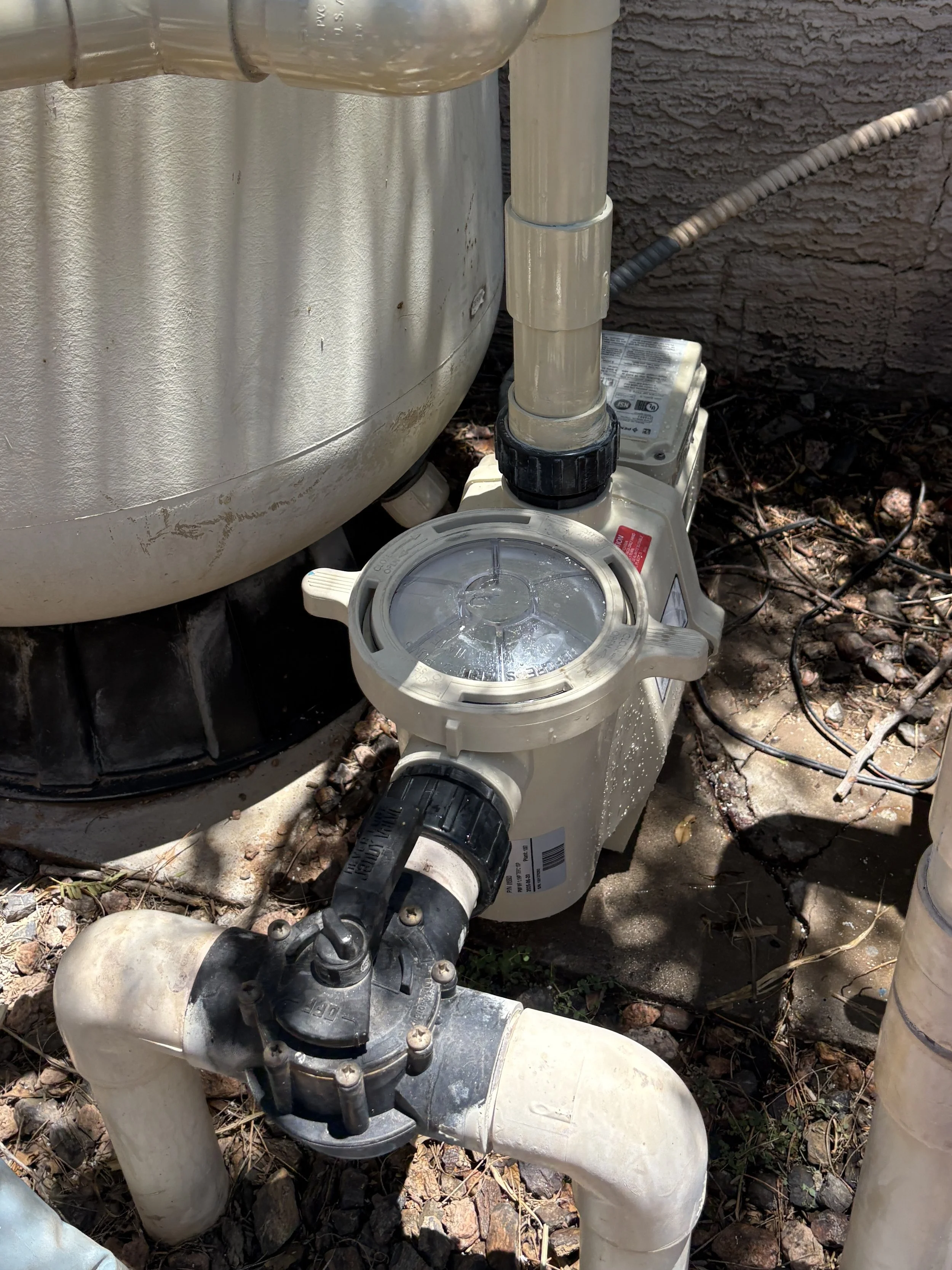 Close-up of an above-ground swimming pool filtration system connected to the pool with PVC pipes, situated outdoors on rocky ground.