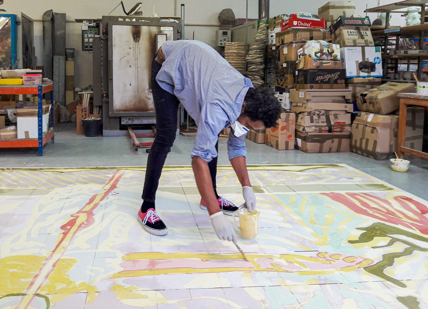Francisco Vidal painting a large mural on the floor inside a workshop with shelves and boxes in the background.