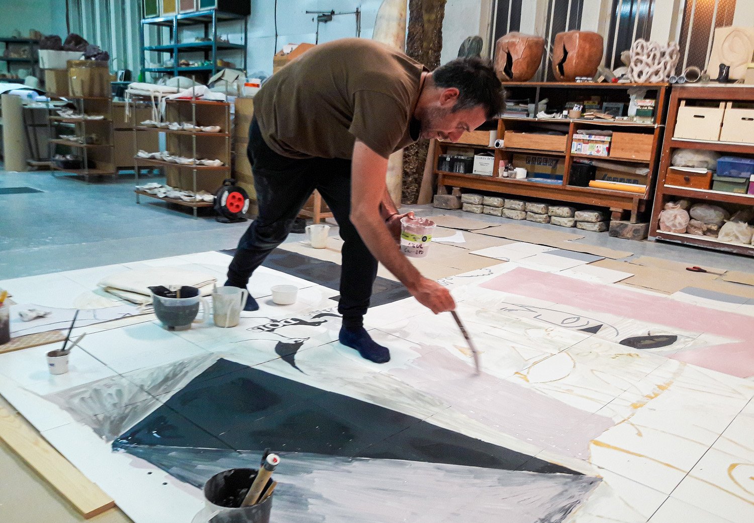 JAS painting a large abstract mural on the floor in an art studio, surrounded by shelves of supplies and artworks.