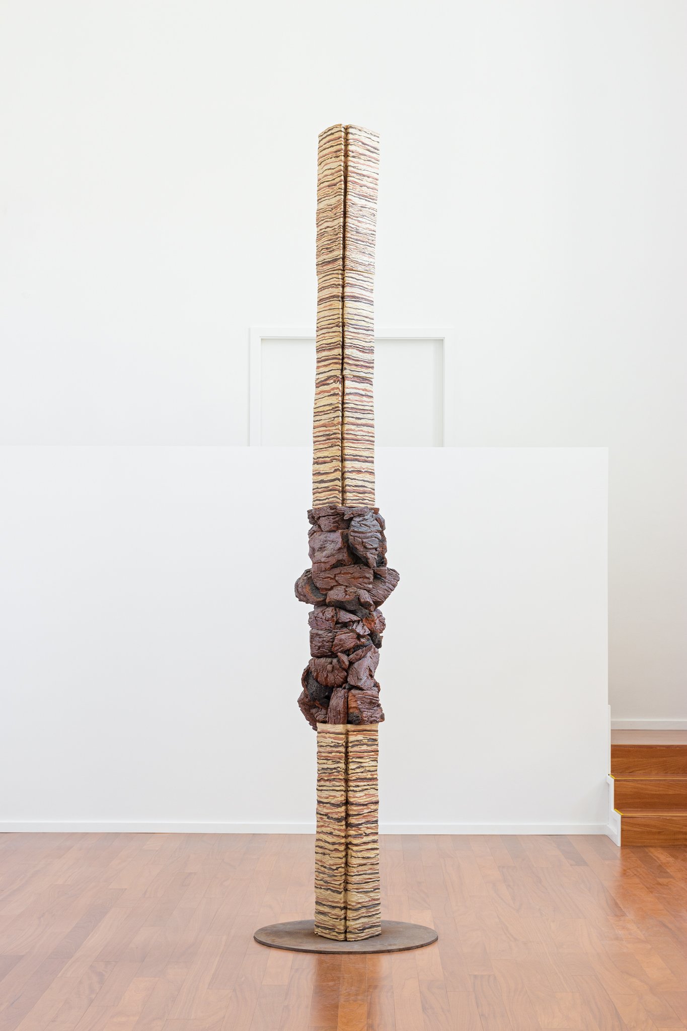 A tall sculpture composed of stacked organic materials, with a rough, irregular base supporting a smooth, striped upper section, displayed in a gallery with white walls and wooden flooring.
