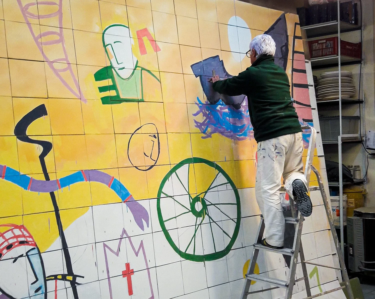 Artist Sobral Centeno creating a colorful mural on a tiled wall, depicting abstract figures, a bicycle wheel, and various shapes and symbols.