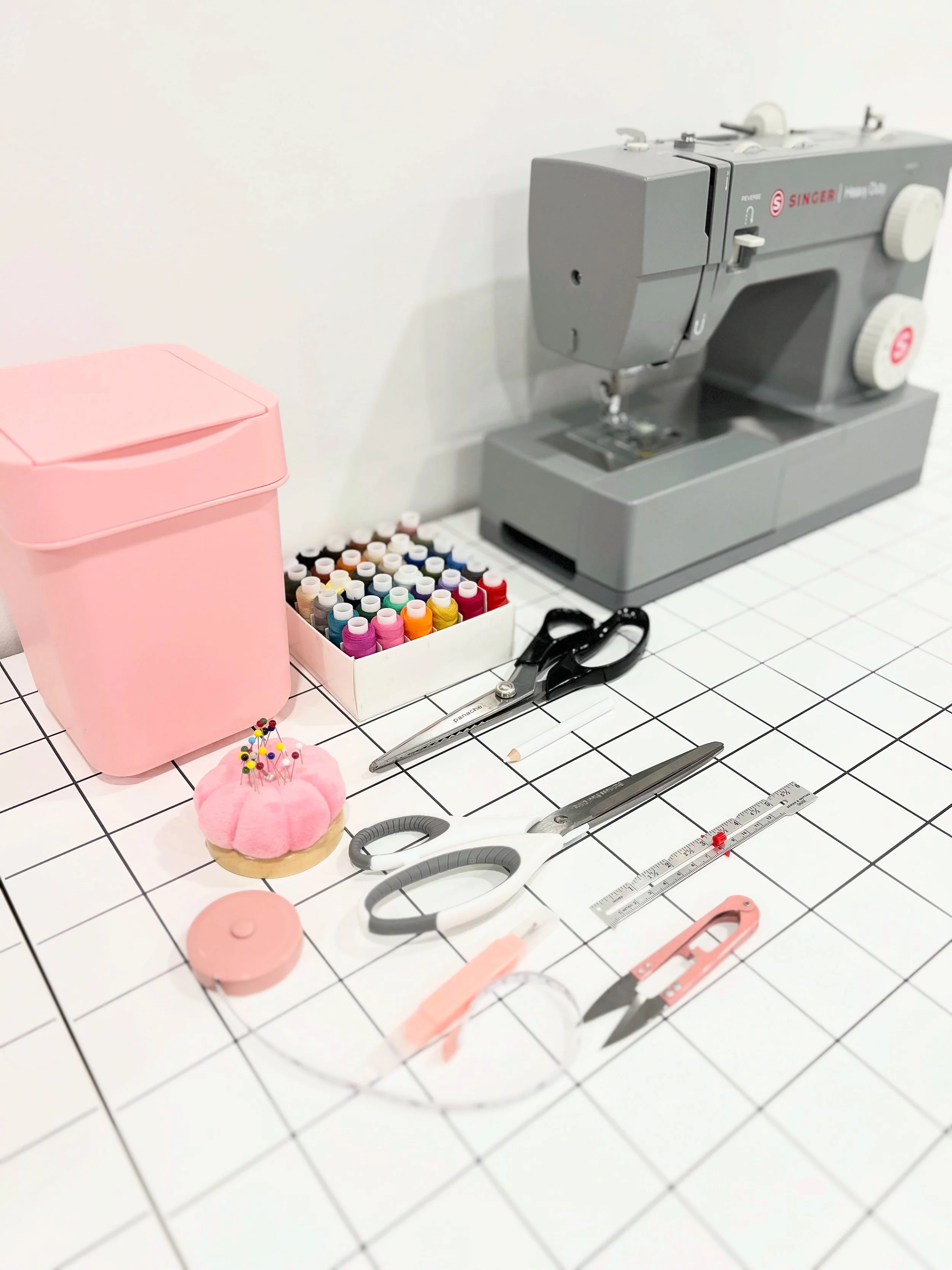 A sewing workspace with a gray sewing machine, a pink trash bin, a box of colorful sewing thread spools, black scissors, a pink pincushion with pins, a ruler, a pair of pink scissors, and other sewing tools on a white tiled surface.