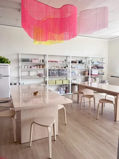 A bright, minimalist workspace with two wooden tables surrounded by matching stools. White shelves on the wall hold various office and art supplies, and colorful paper lanterns hang from the ceiling for decoration.