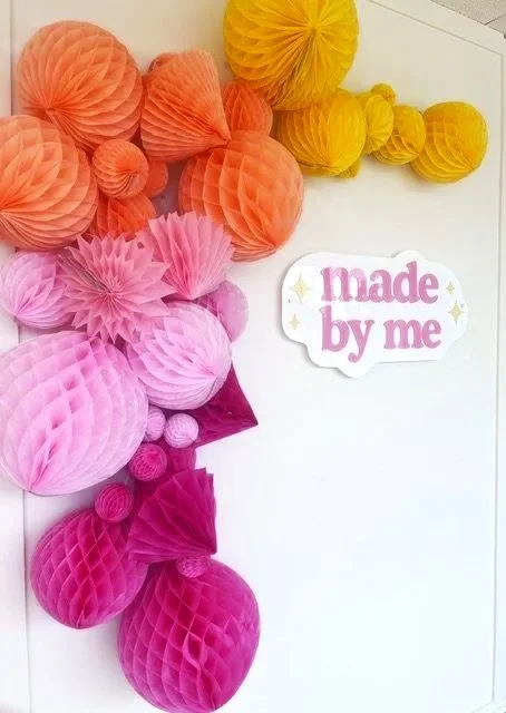 Colorful paper honeycomb balls in shades of orange, yellow, pink, and magenta hanging on a white wall with a "Made by me" sticker.