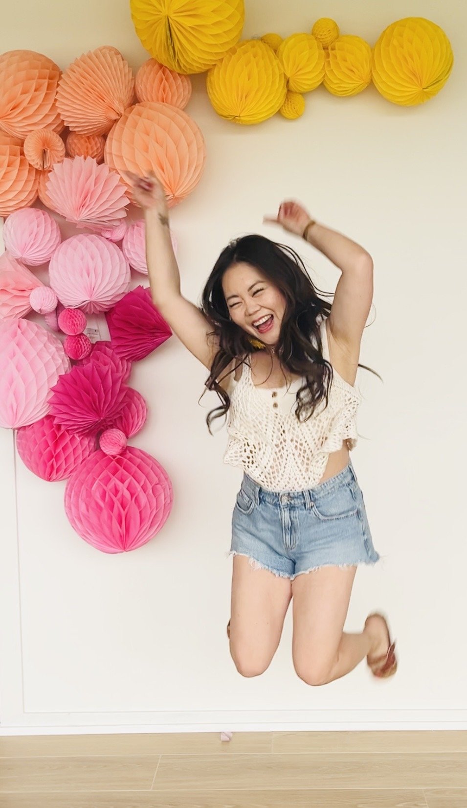 A woman jumping and smiling in front of a colorful paper decoration wall with yellow, pink, and peach honeycomb balls.