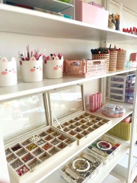 Organization shelf with various stationery supplies, including decorated cups with bunny faces, small baskets with pens and pencils, and a jewelry box.