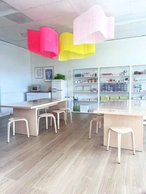 Modern room with white walls, colorful cloud-shaped pendant lights, white shelving with various items, a white fridge, and a wooden table with matching stools.