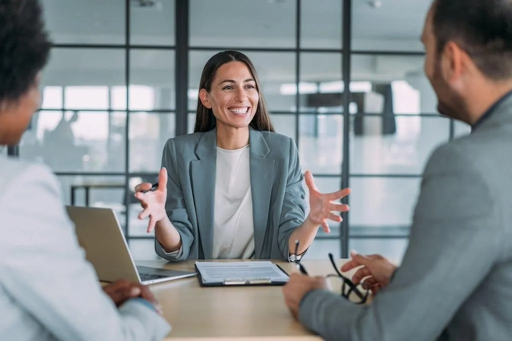 A woman in a light blue blazer smiling and gesturing with her hands during a business meeting with two men in a modern office.