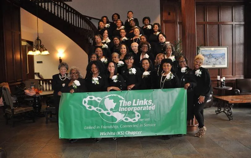 Group of women holding a green banner that reads 'The Links, Incorporated, Wichita (KS) Chapter,' inside a wood-paneled room with a staircase, painting, and table.