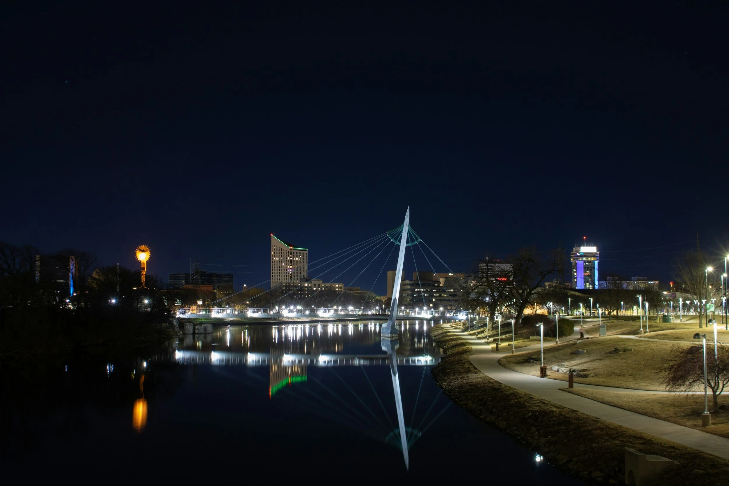 Nighttime cityscape of Wichita, KS, featuring illuminated modern buildings, a unique bridge with cables, and their reflections on a calm river, with a dark sky overhead.