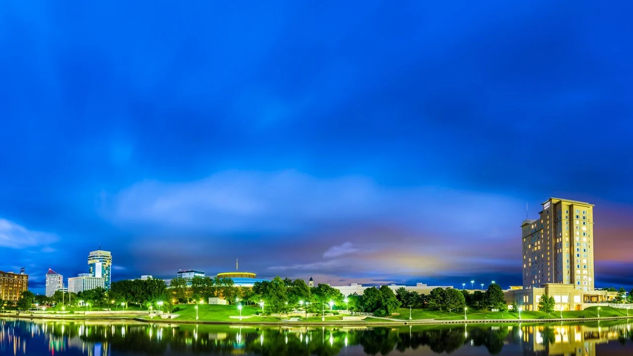 Nighttime Wichita, KS, cityscape with illuminated buildings, trees, and a water reflection.