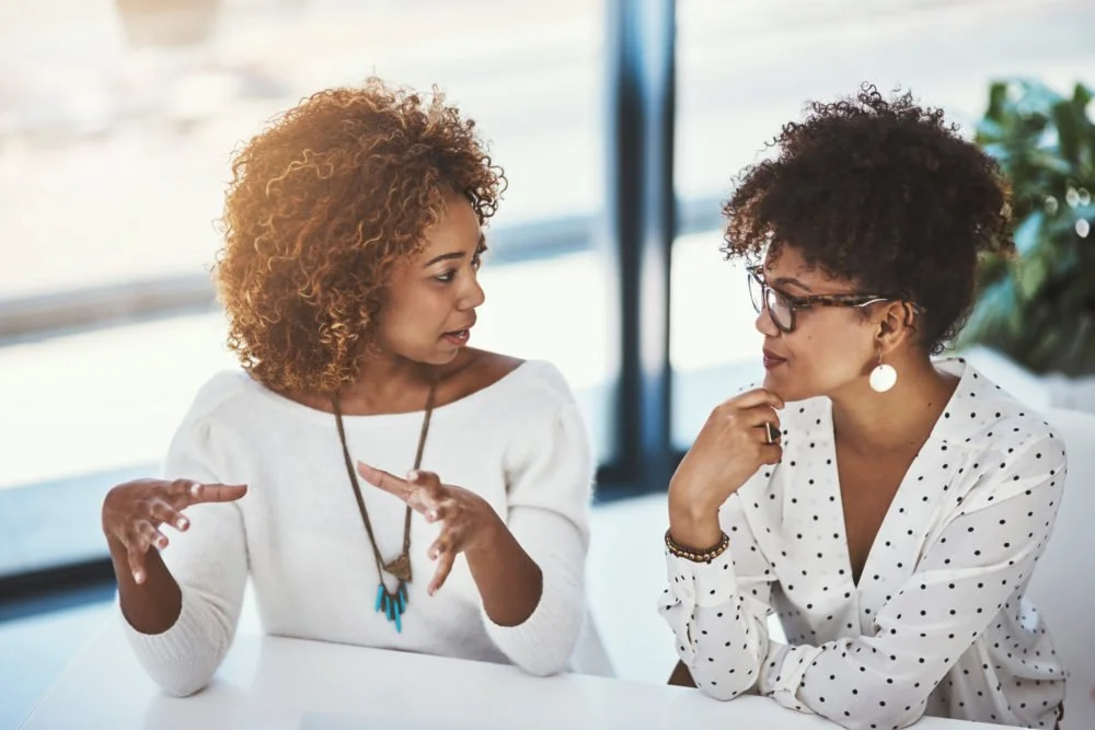 Two women of African descent in conversation at a table indoors, one gesturing with hands and the other listening thoughtfully.
