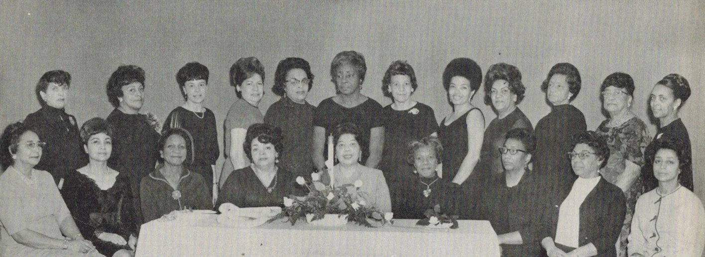 A group of women and girls posed together for a photo, with some seated at a table and others standing behind them. The setting appears to be indoors against a plain wall.