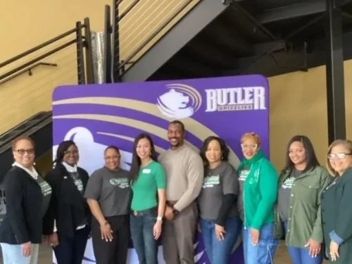 Group of women and one man standing in front of a purple Butler University backdrop, smiling indoors.