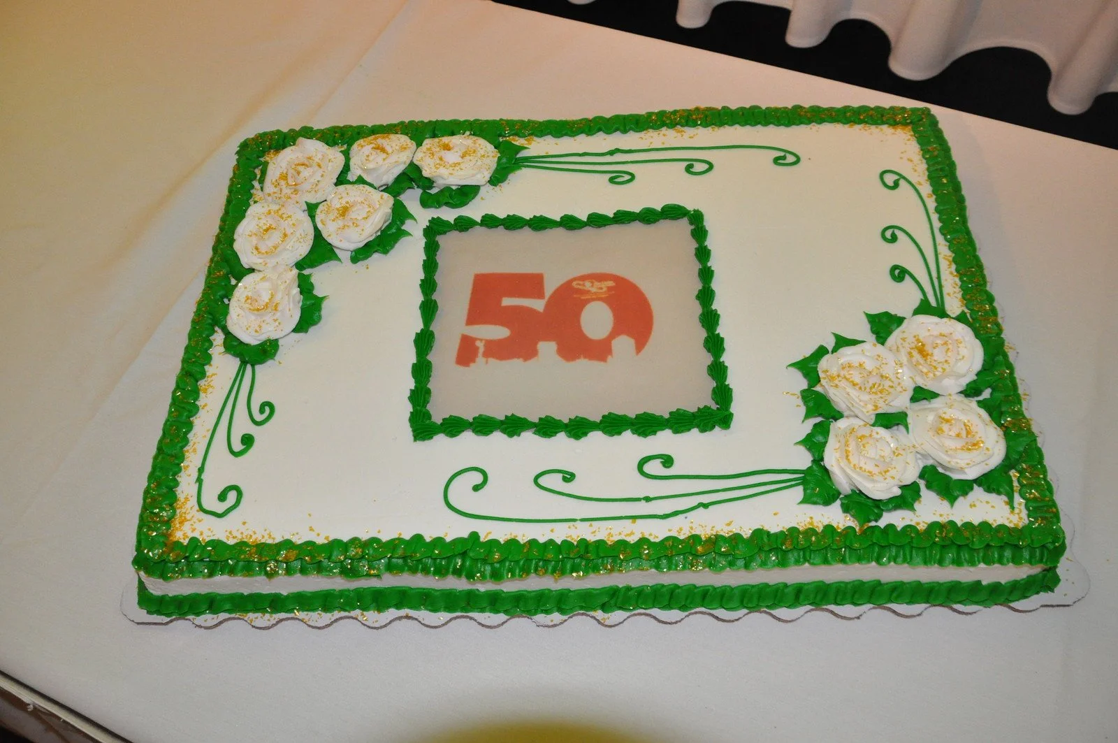 Square white cake decorated with green icing border and white rose-like flowers with gold sprinkles, featuring a red "50" in the center.