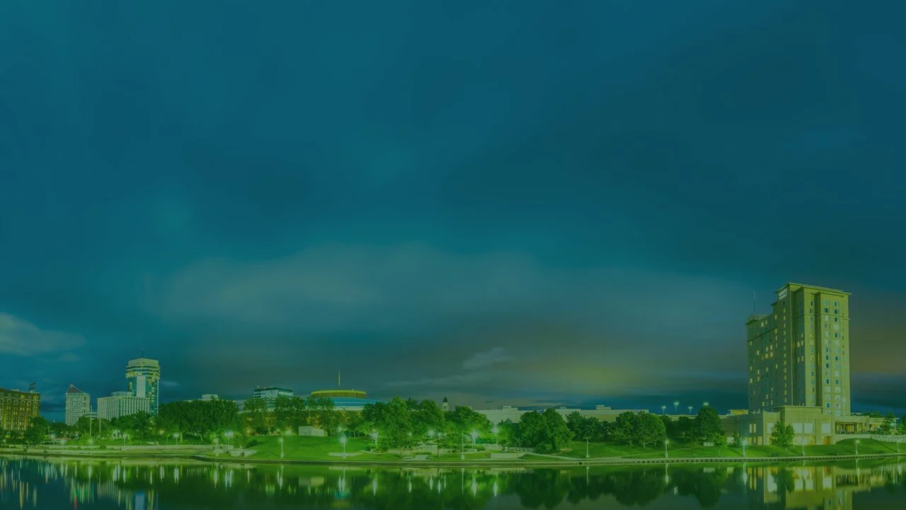 Wichita, KS, city skyline with multiple tall buildings and a large body of water in the foreground, under a cloudy sky.