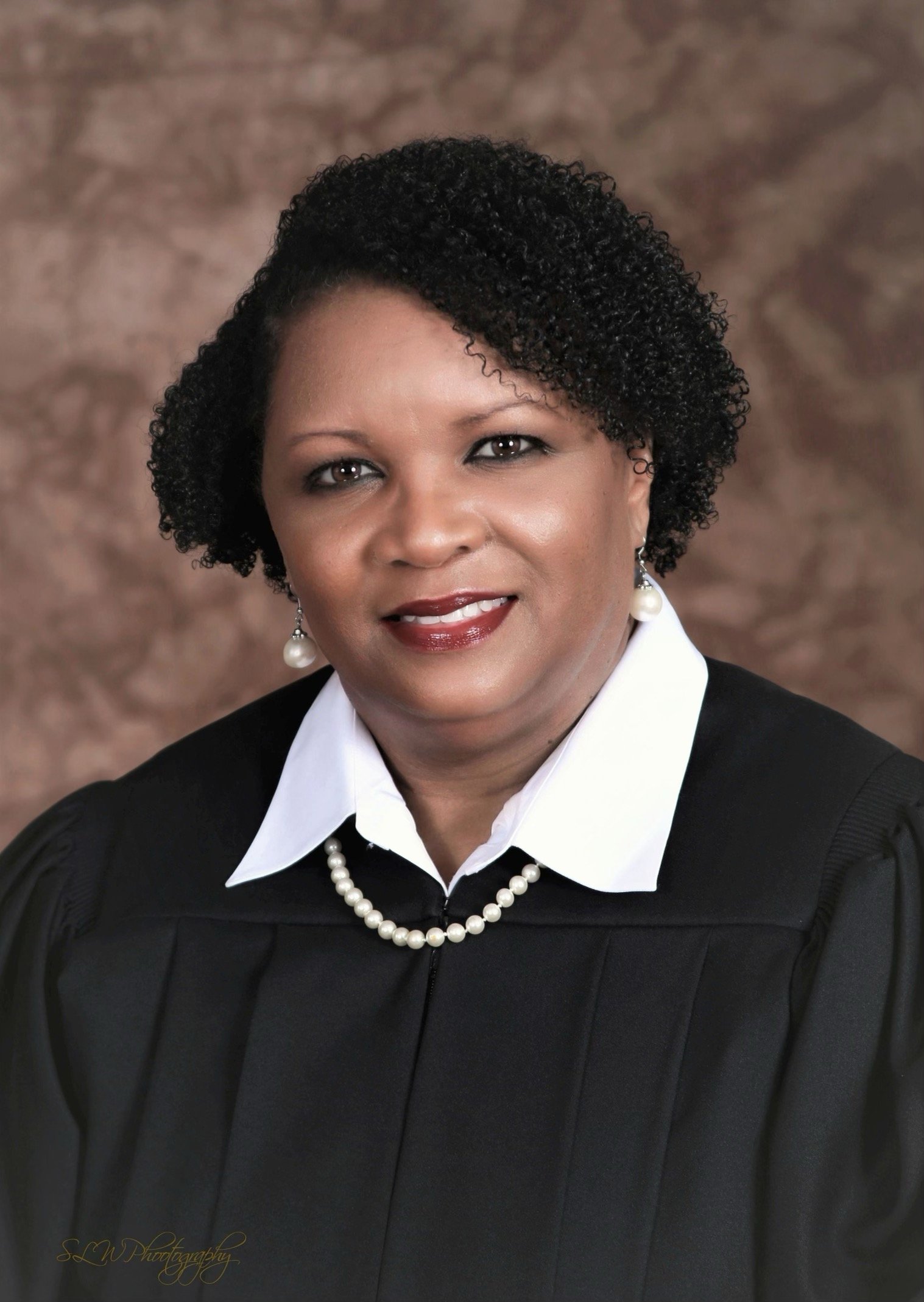 A woman with short curly black hair, wearing a black judicial robe over a white shirt, pearl necklace, and pearl earrings, smiling against a brown textured background.