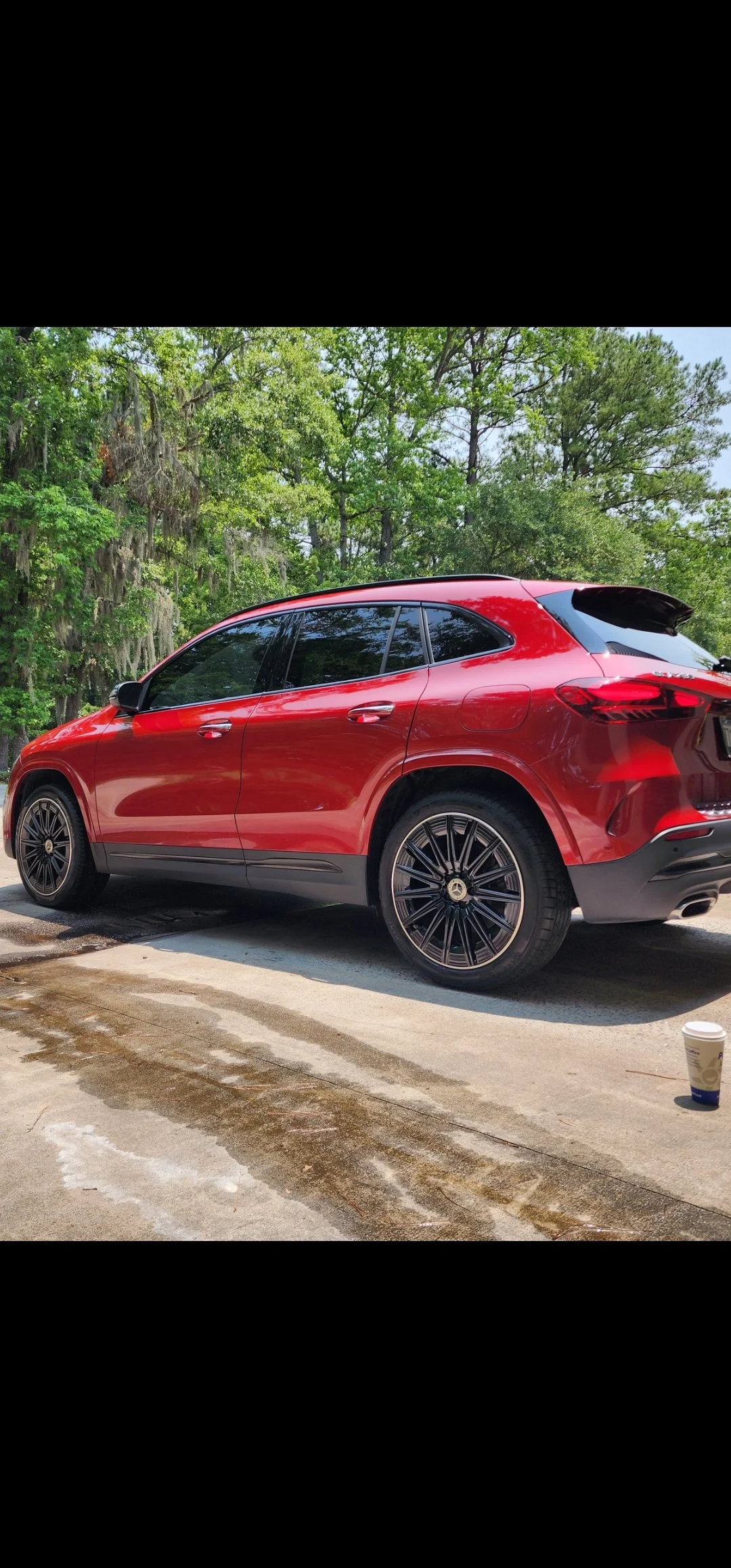 Red Mercedes-Benz SUV parked outdoors on wet concrete, with green trees in the background and a disposable coffee cup on the ground.