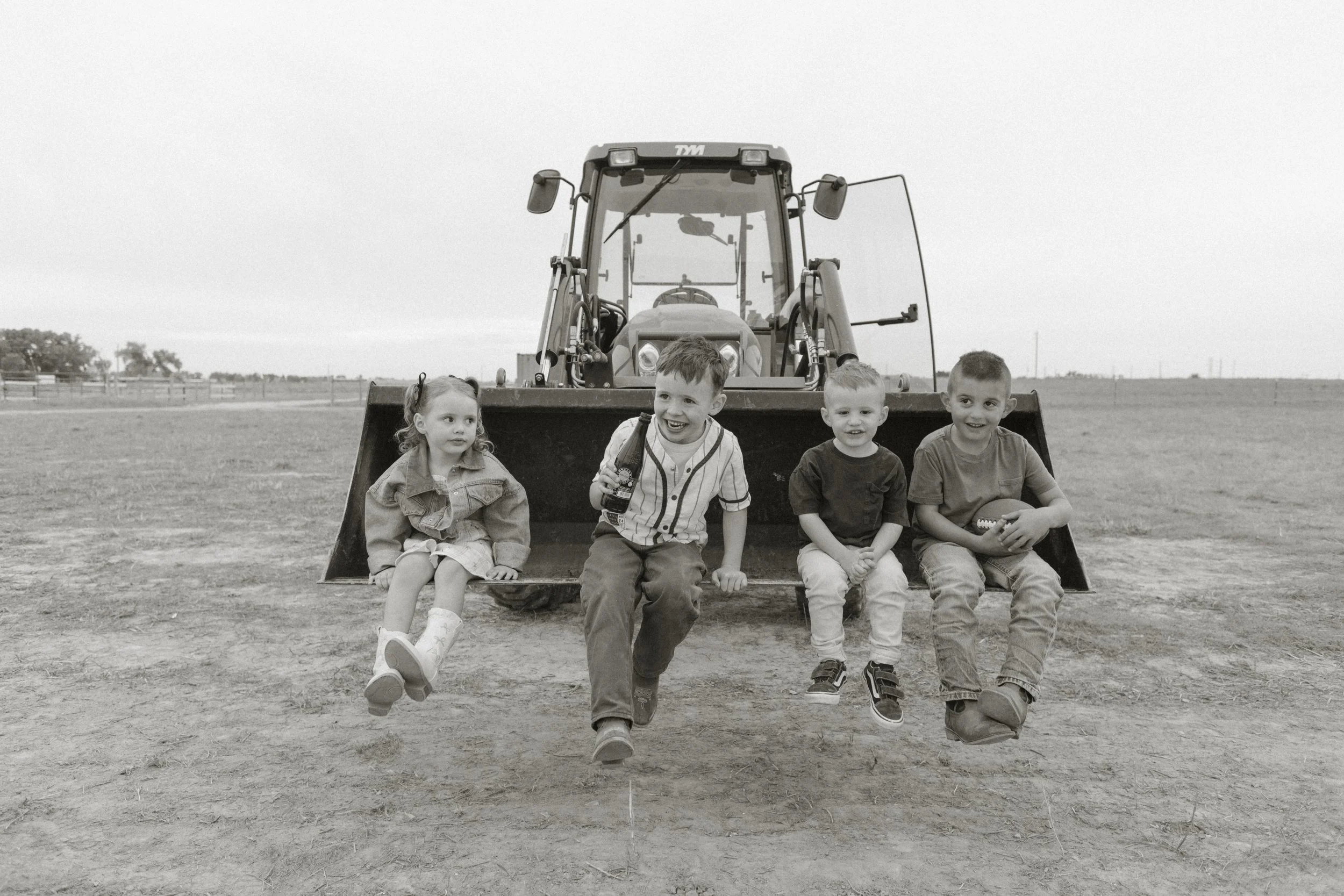 Four children sitting on the front of a large tractor on a field, with a cloudy sky and open landscape in the background.
